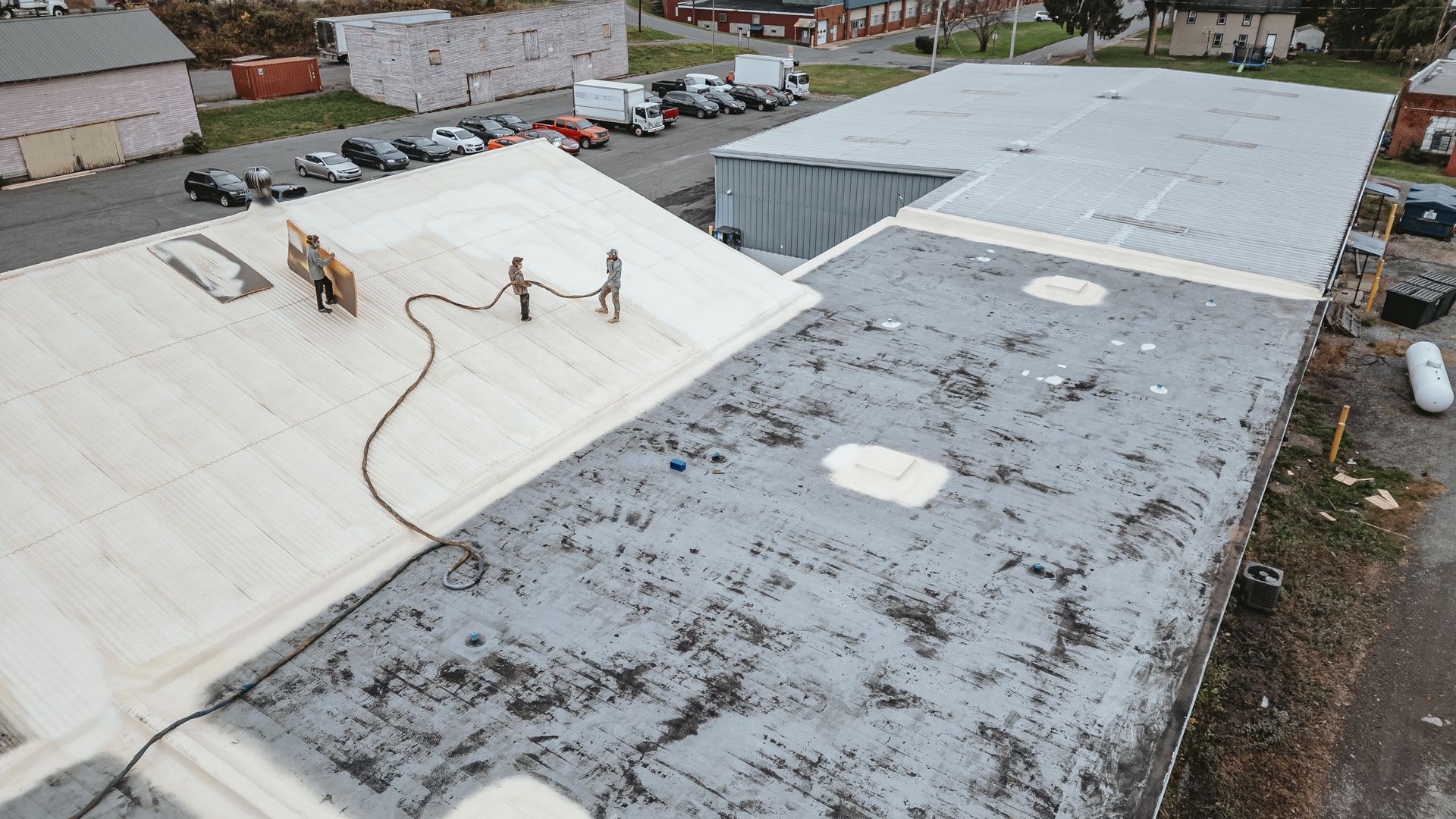 Workers applying coating to a commercial roof. Grey and white roof sections, parked vehicles in the background.