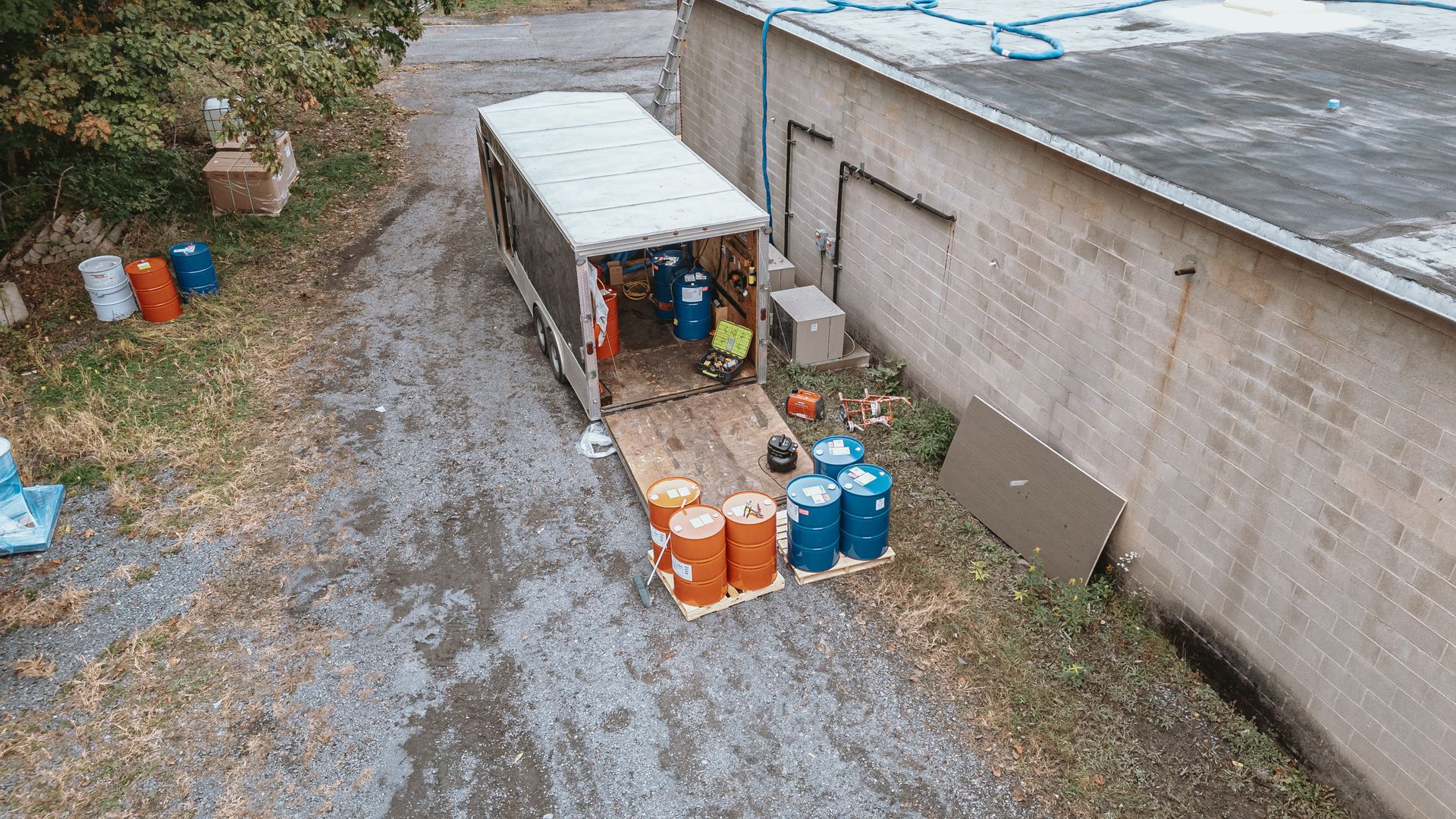 A trailer is open, filled with blue and orange drums. Located on gravel next to a brick building.