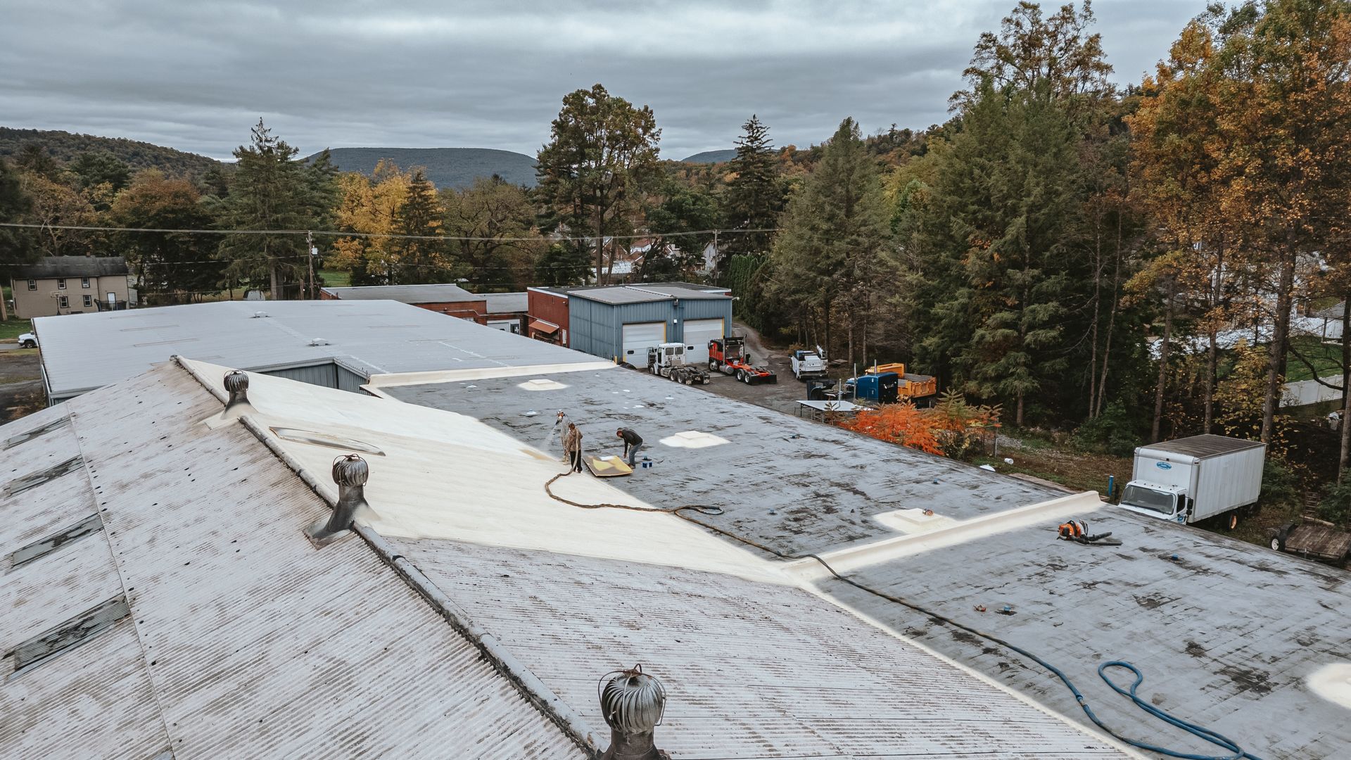 Workers on a large flat roof, installing insulation. Surrounding trees and buildings in the background. Overcast sky.