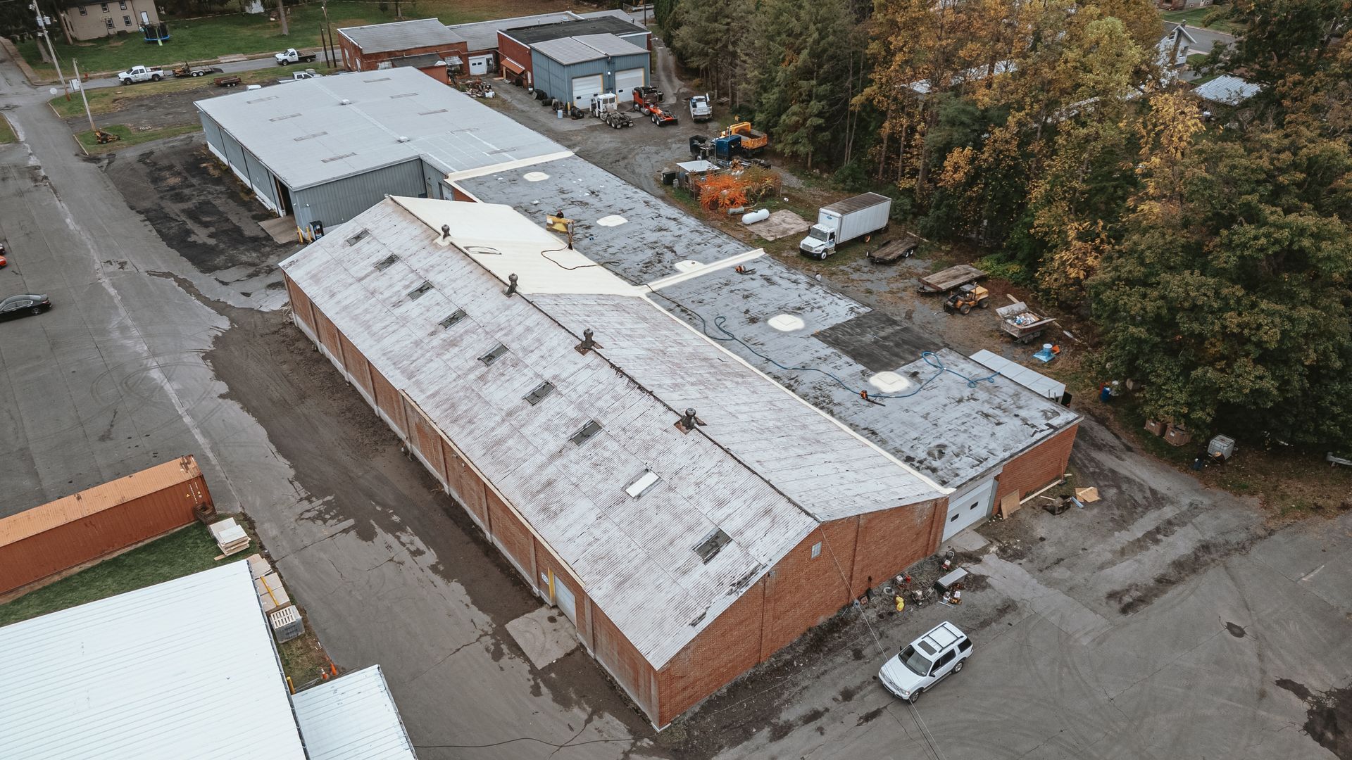 Aerial view of industrial buildings with gray roofs and a parking area, surrounded by trees.