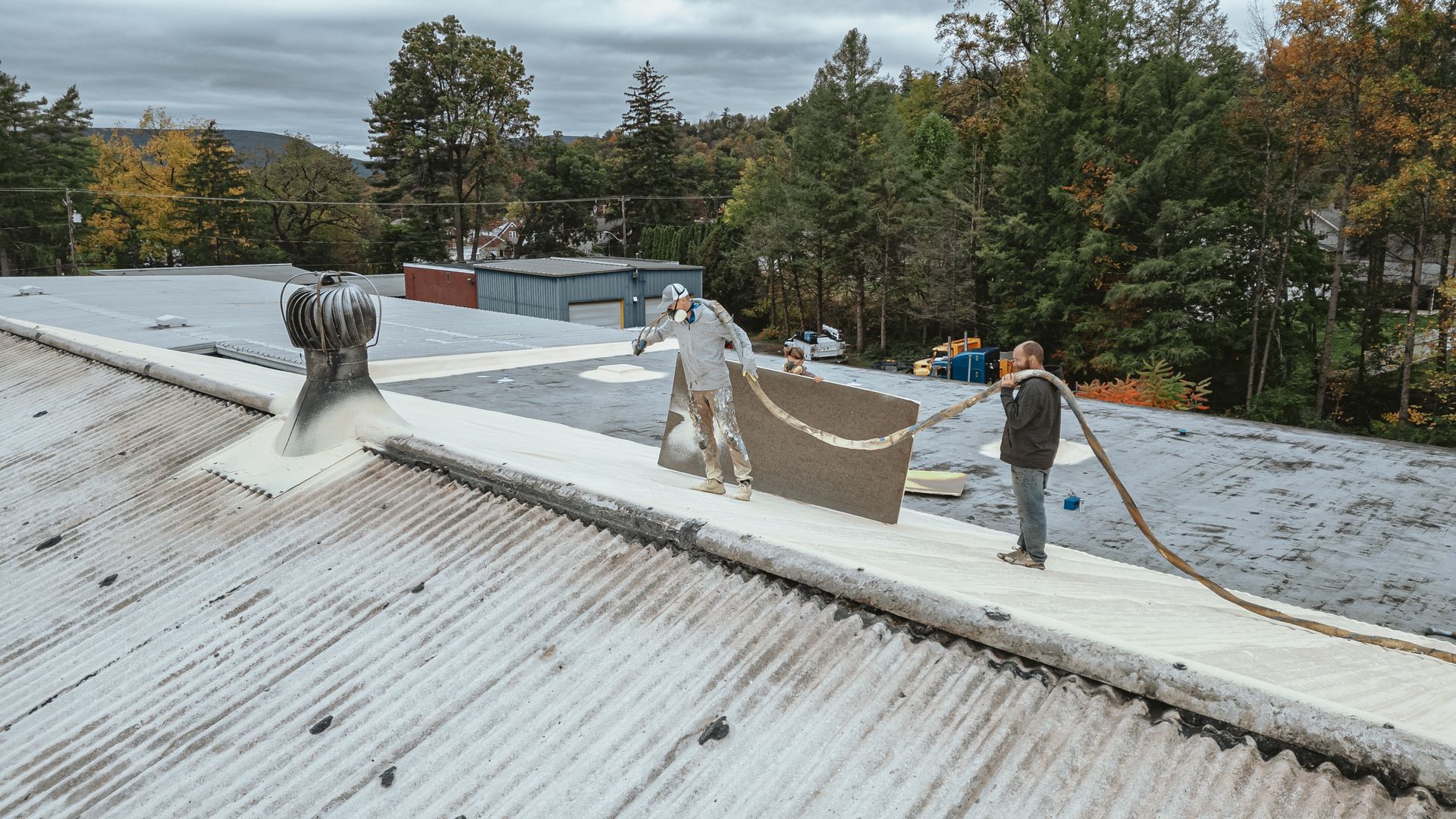 Two people on a roof applying white spray, likely roofing work, with trees and a cloudy sky in the background.