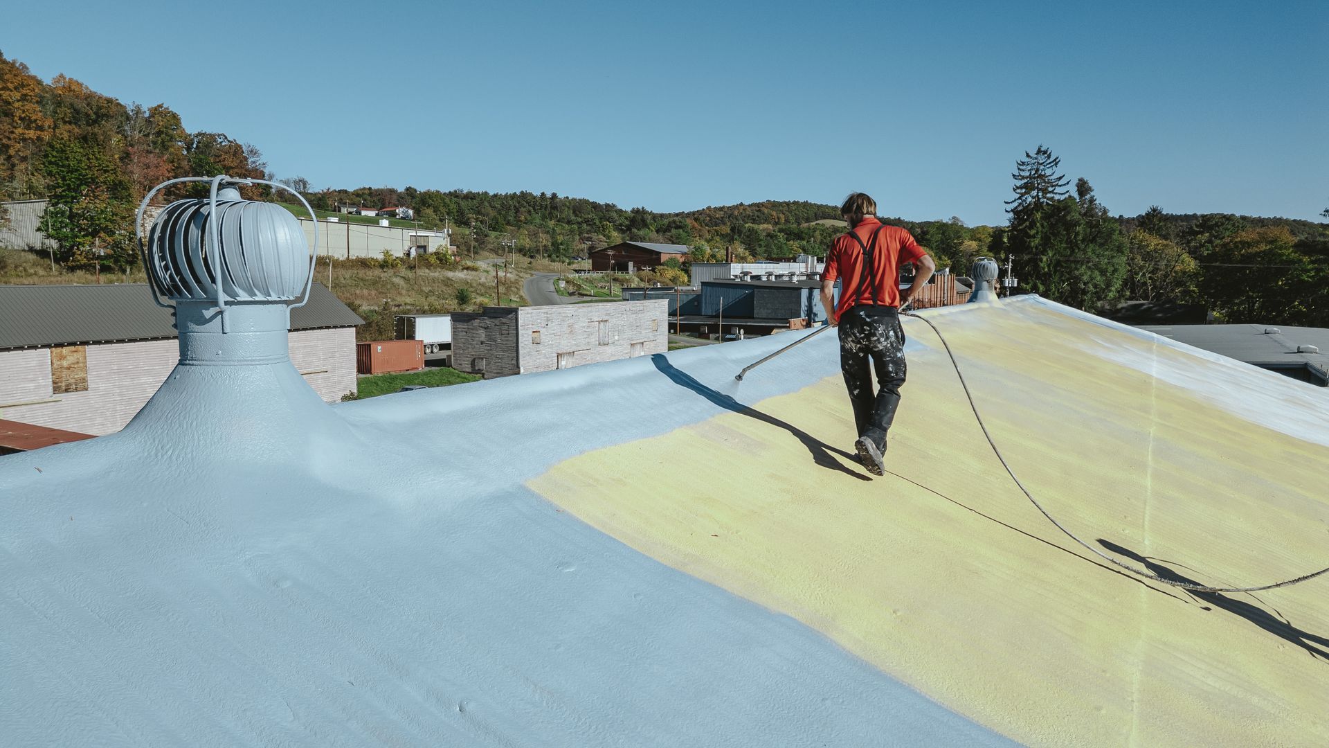 Man walks on painted rooftop, with blue and yellow sections. Trees and buildings in the background.
