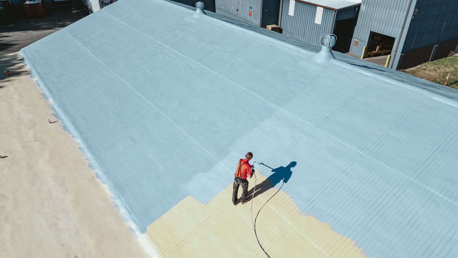 Person in orange spraying light blue coating on a corrugated metal roof.