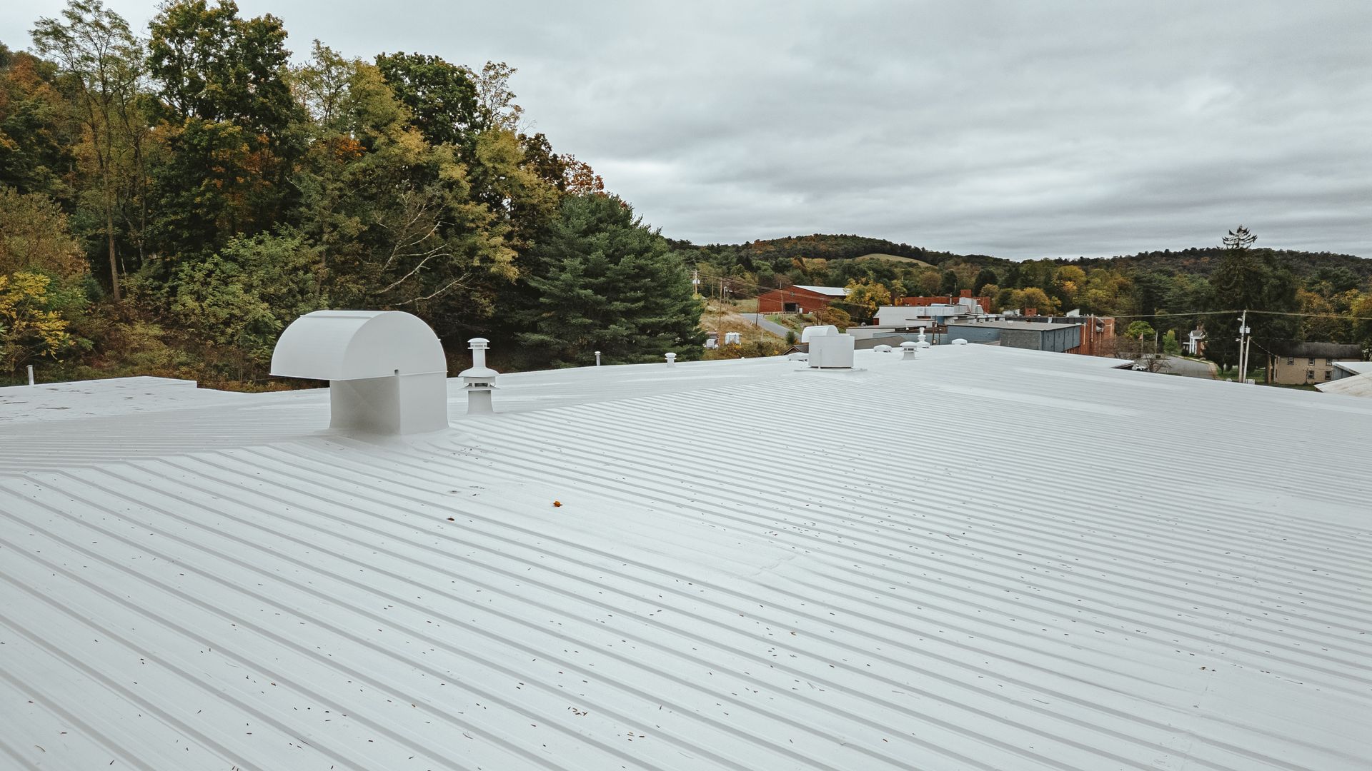 White commercial roof with ventilation equipment, trees, and buildings in the background under a cloudy sky.