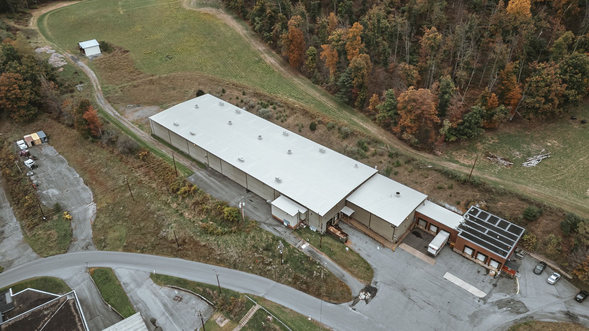Aerial view of a long, low industrial building with a corrugated metal roof. Located near a wooded area with parking.