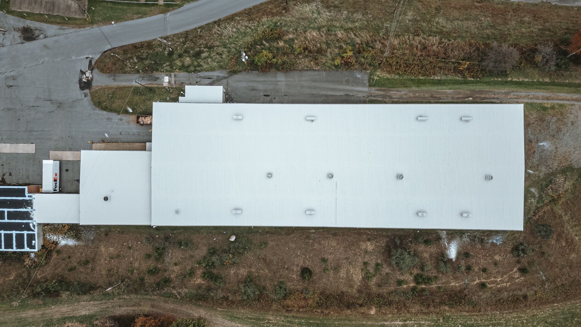 Aerial view of a rectangular industrial building with a white roof and surrounding patches of grass and asphalt.