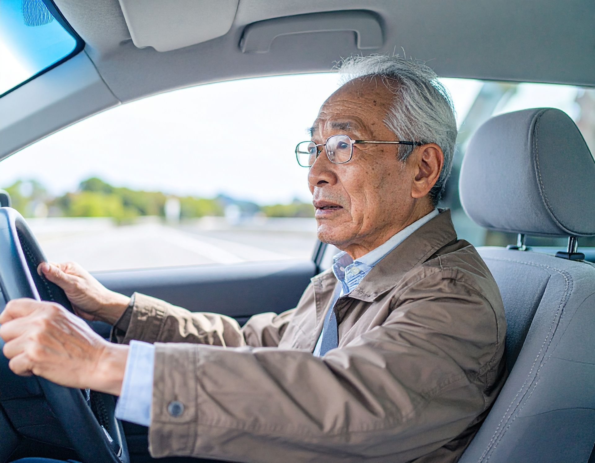An older person with gray hair and glasses sits in the driver's seat of a car, hands on the steering wheel.