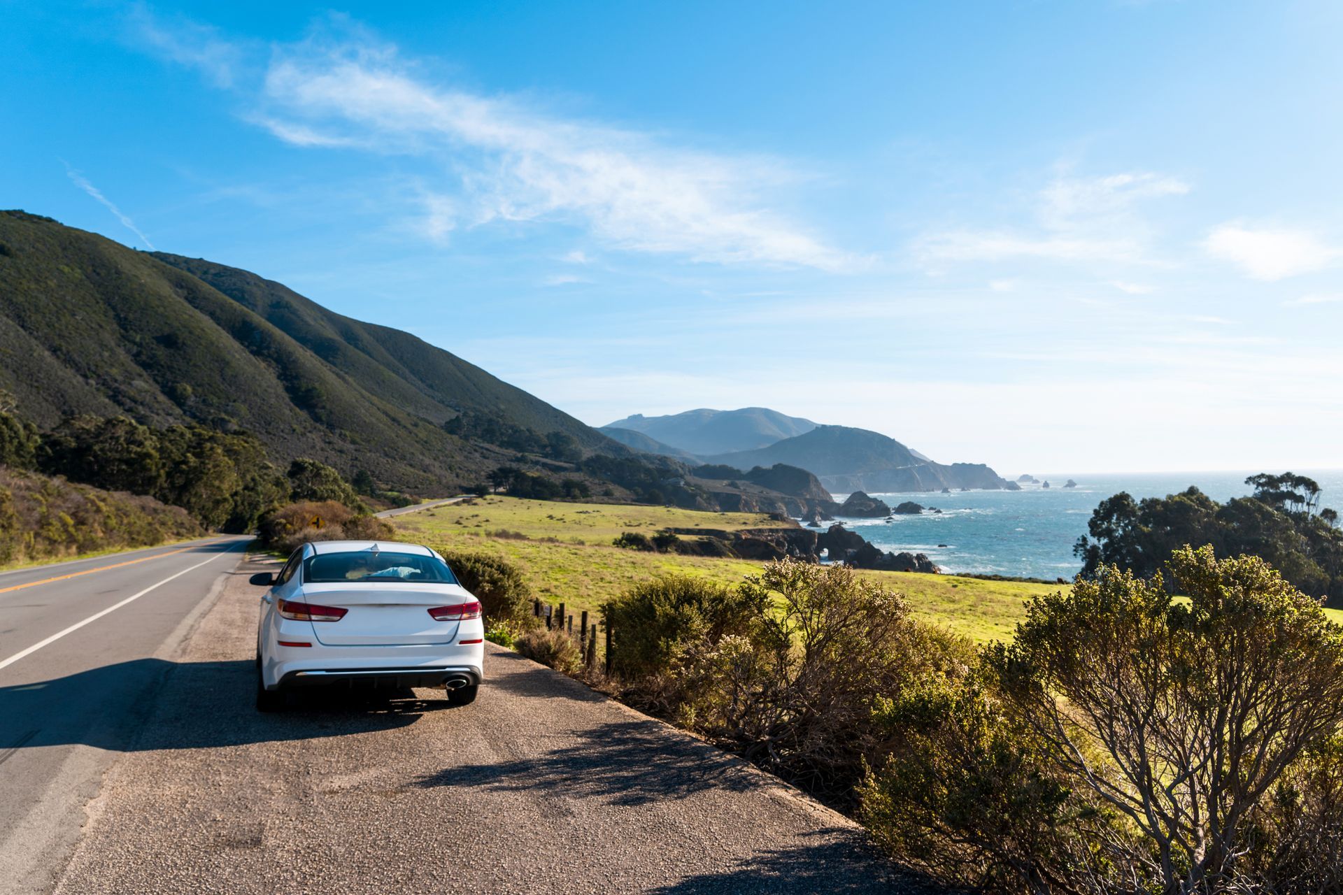 White car parked on a scenic coastal road with mountains, ocean, and blue sky.