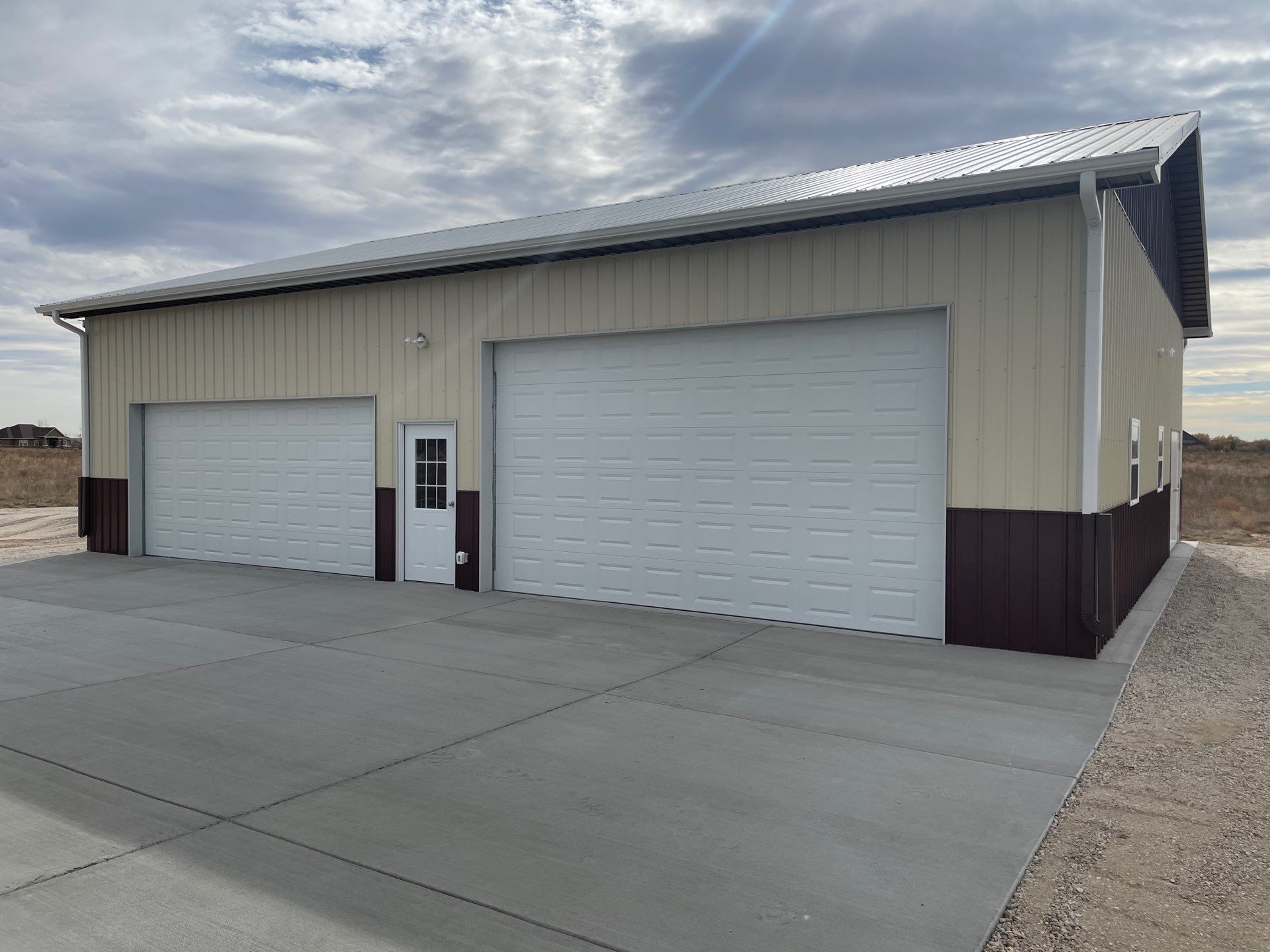 Two-door metal garage building with white roll-up doors and a concrete apron under a cloudy sky