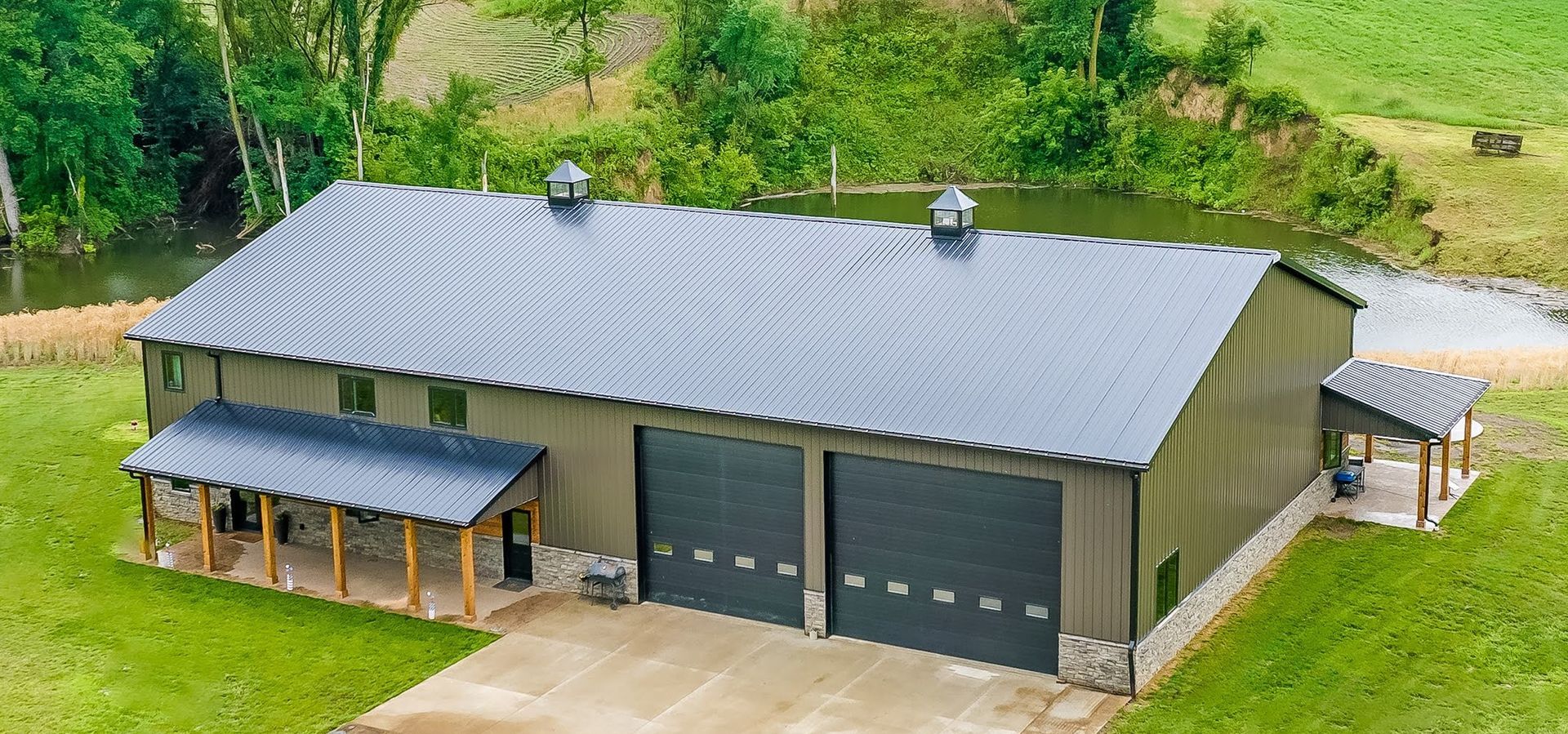 A large, olive-green metal building with a dark gray roof. It has two garage doors and a porch with stone accents.