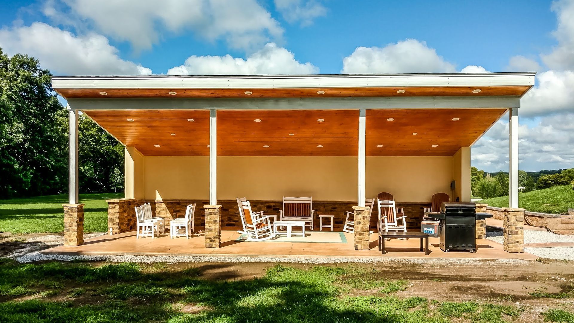 Pavilion with seating, a grill, and light-colored walls and pillars, set against a blue sky and green grass.