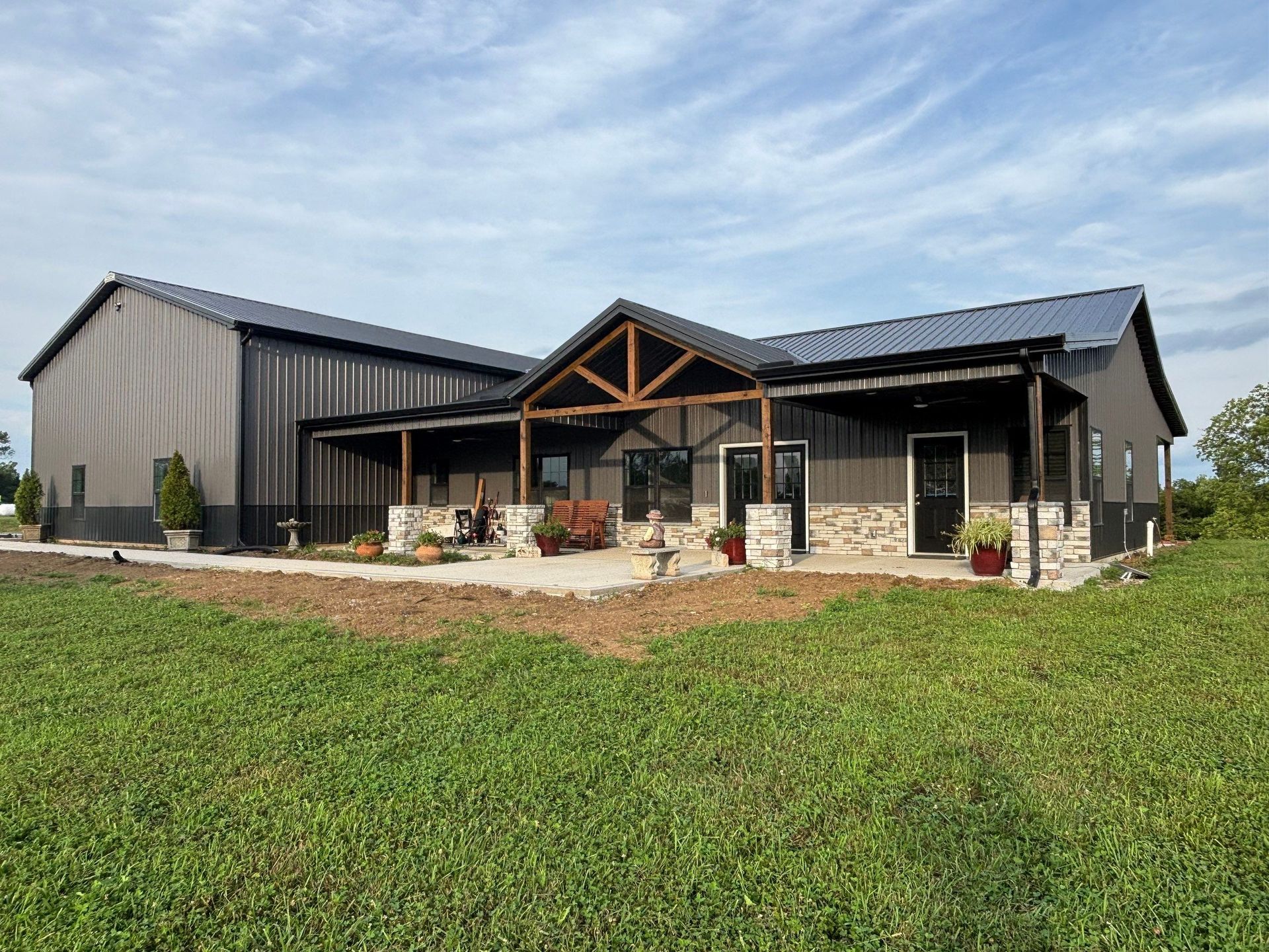Modern gray metal barn-style house with porch, in a grassy field under a blue sky.