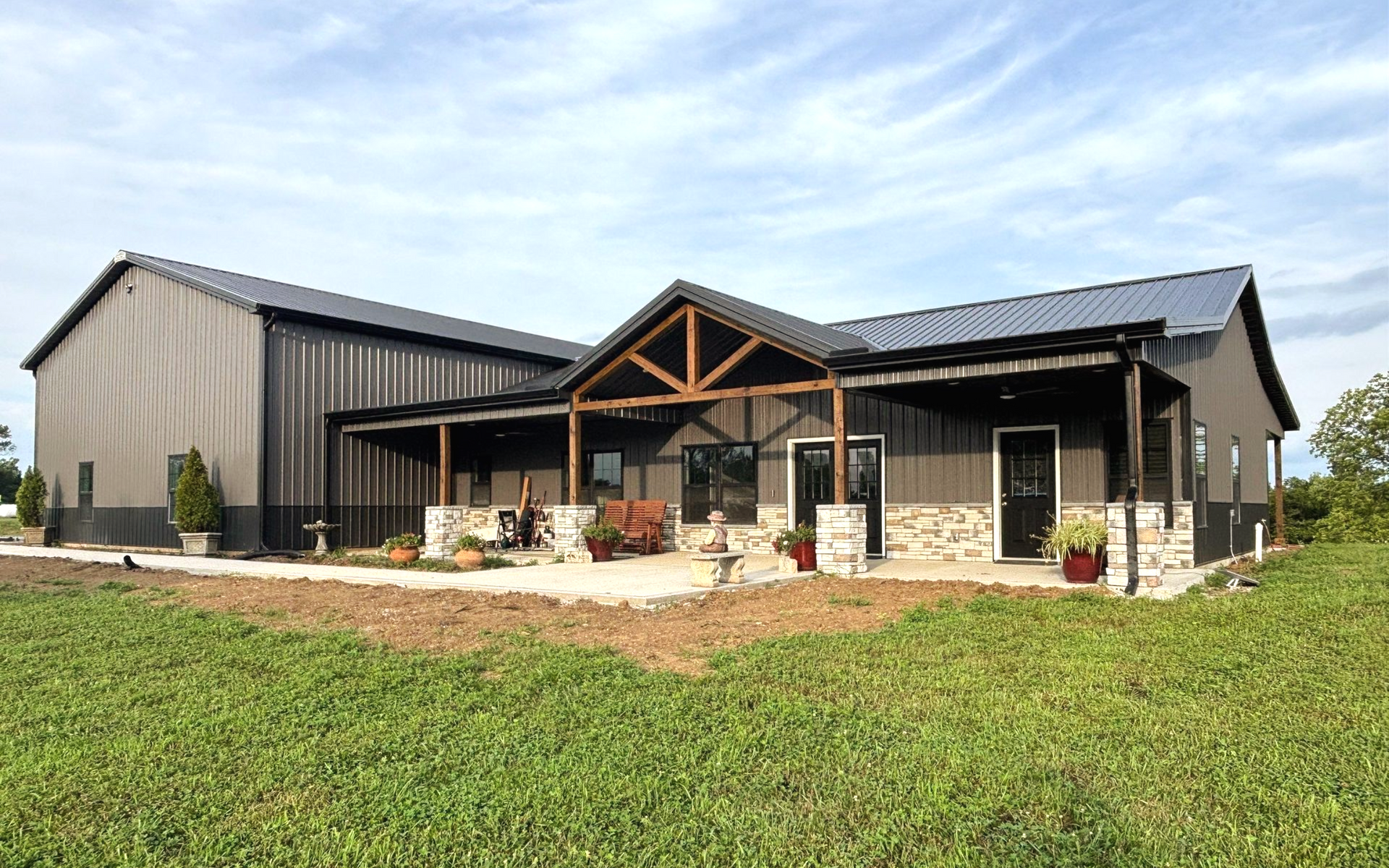 Barn-style building with a porch and metal roof. Dark gray siding, stone accents, and green lawn.