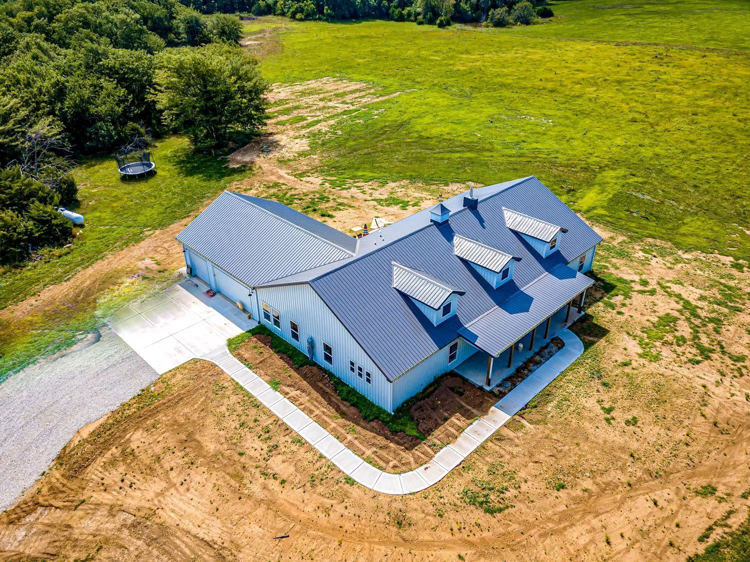 Aerial view of a light blue house with a gray metal roof, dormers, and a concrete driveway on a grassy landscape.