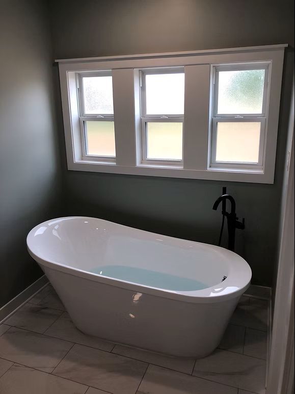 Freestanding white bathtub filled with water under three small frosted windows. Green walls, gray tile floor.