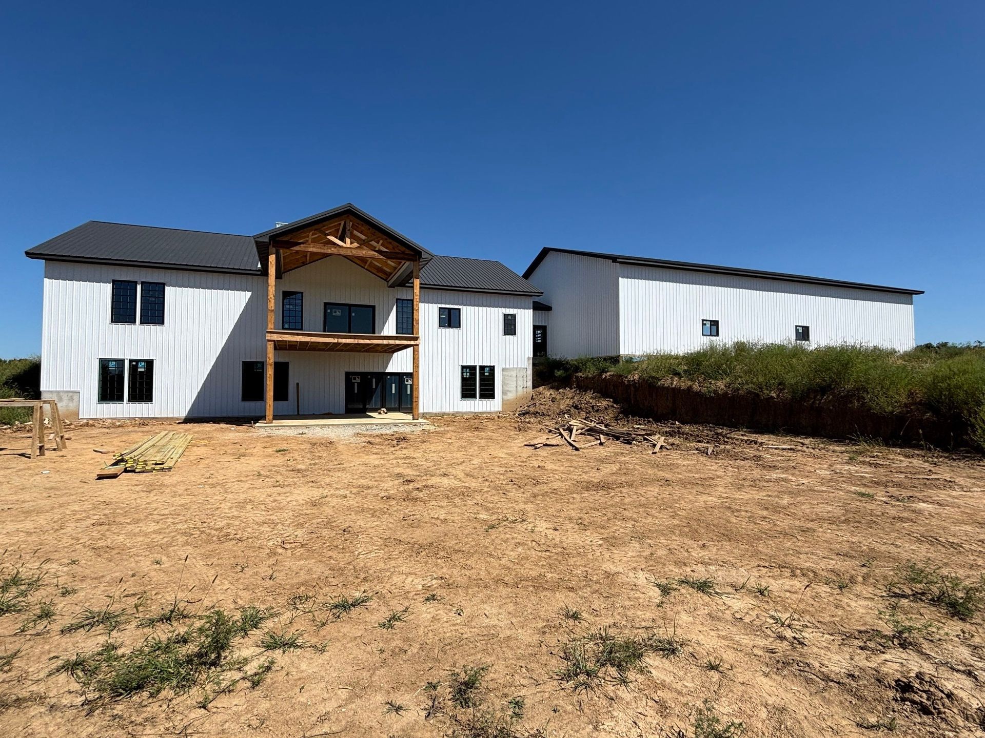 White house under construction with a black roof and a detached building on a dirt lot against a blue sky.