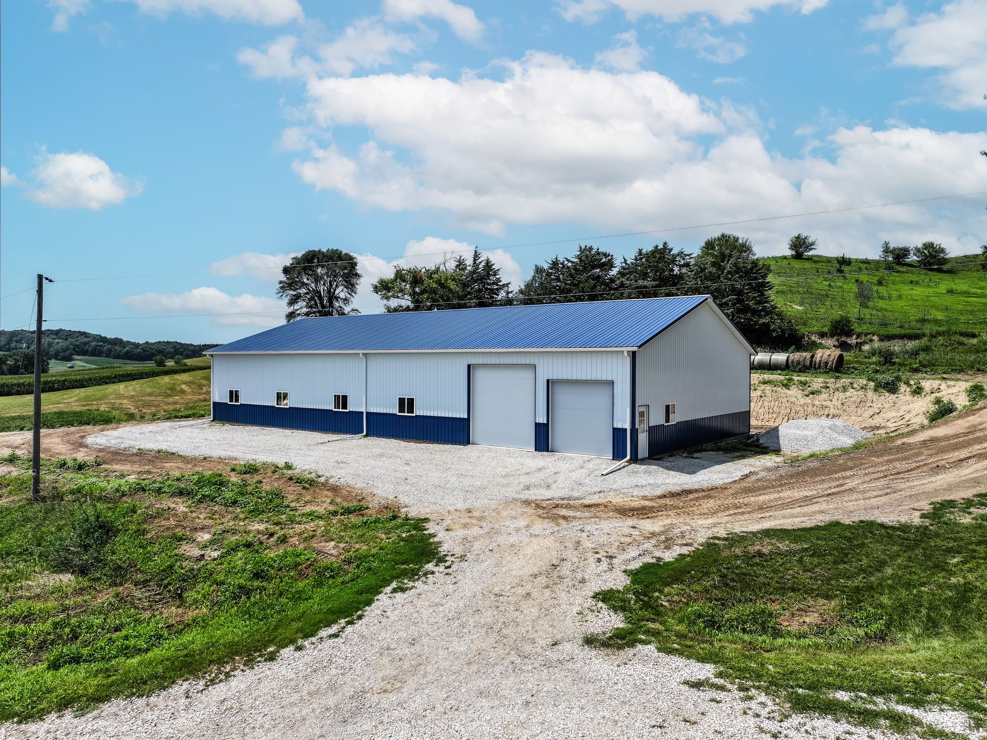Blue and white metal building with two garage doors on gravel driveway, rural setting.