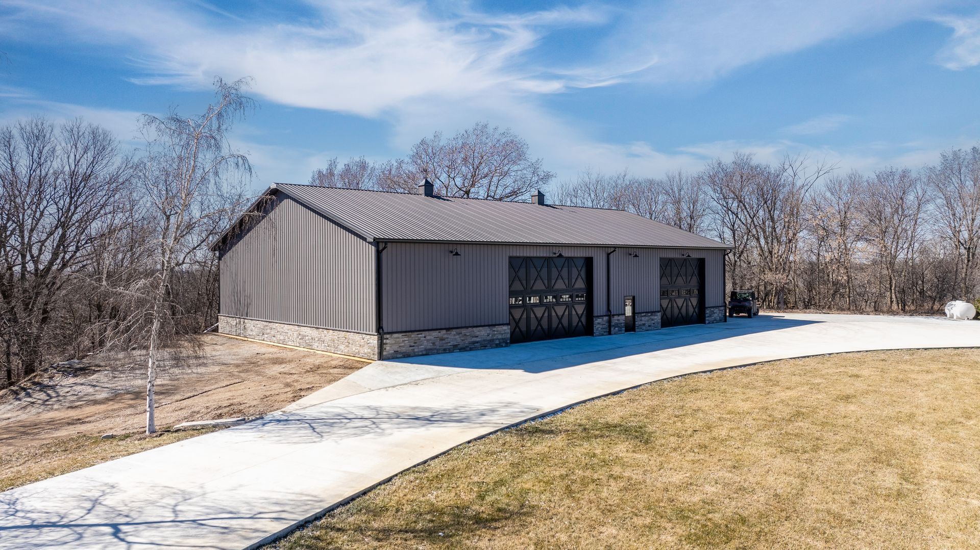 Gray metal building with two garage doors, surrounded by trees and a curved concrete driveway.