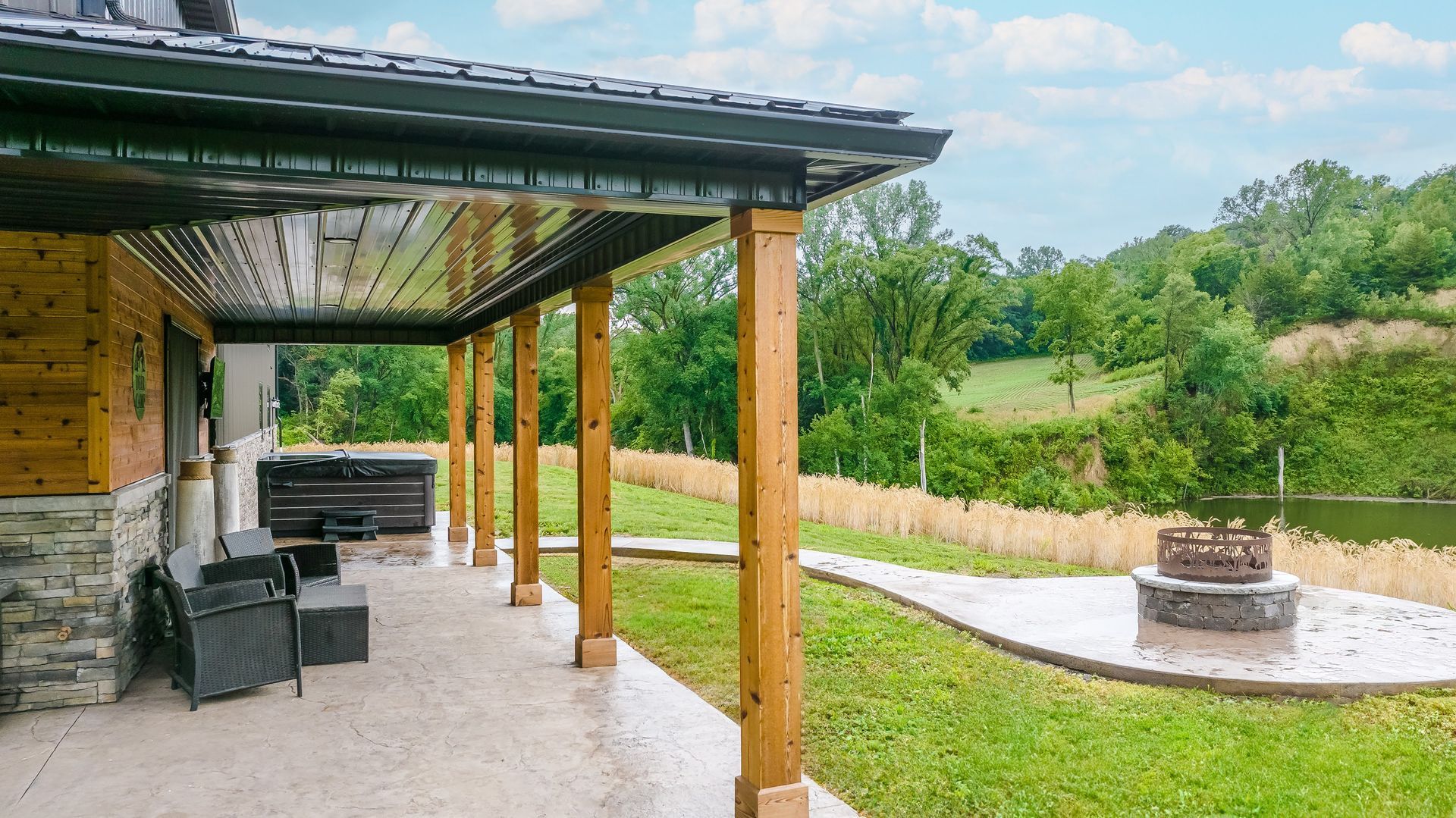 Covered patio with wooden posts, overlooking a green lawn, fire pit, and a lake in the background.