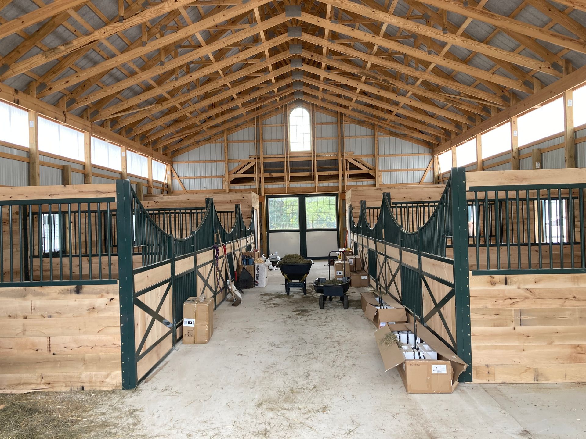 Interior view of a barn with horse stalls, light wood, green accents, and a central aisle.