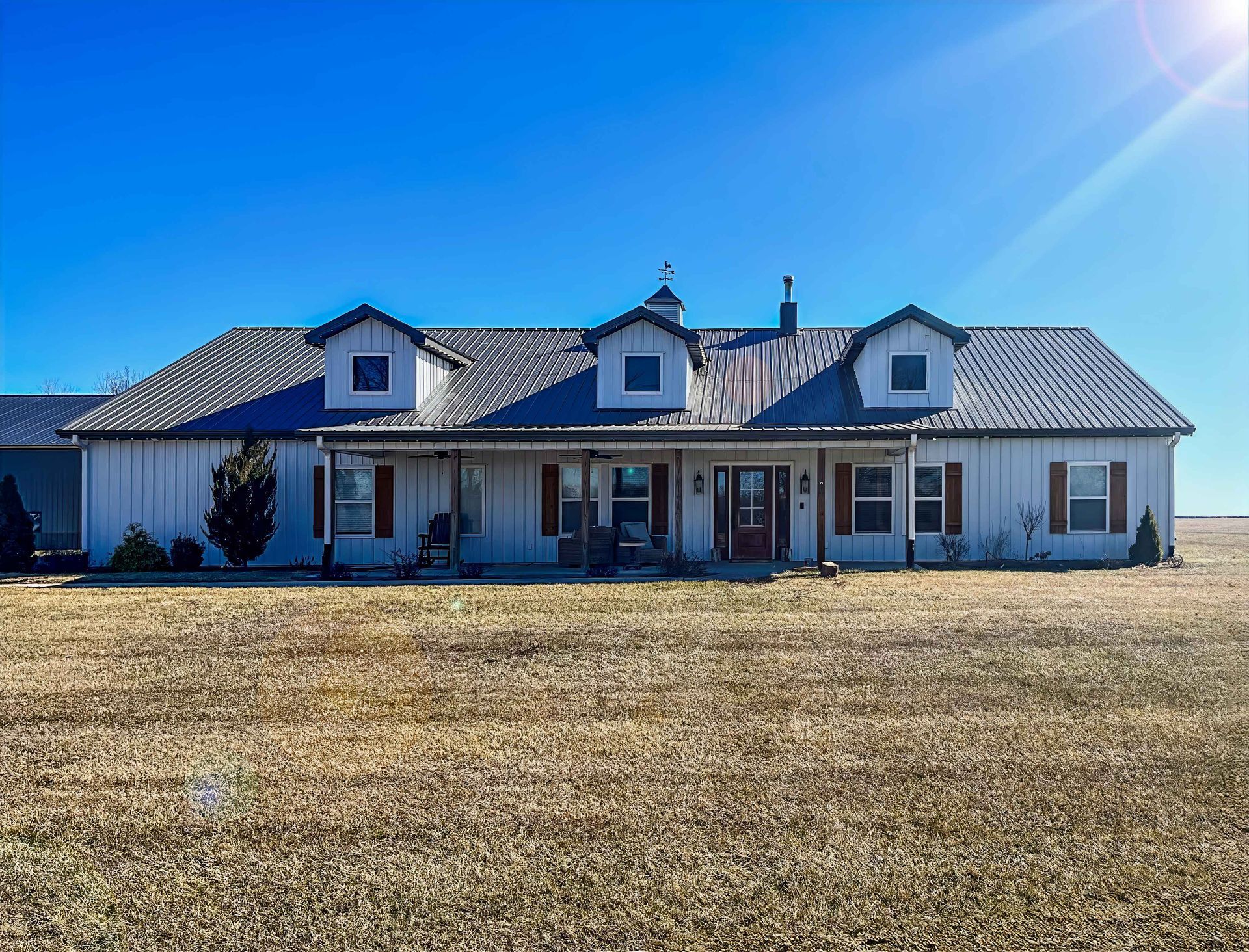 White farmhouse with metal roof, brown shutters, front porch, set in a field under a bright blue sky.