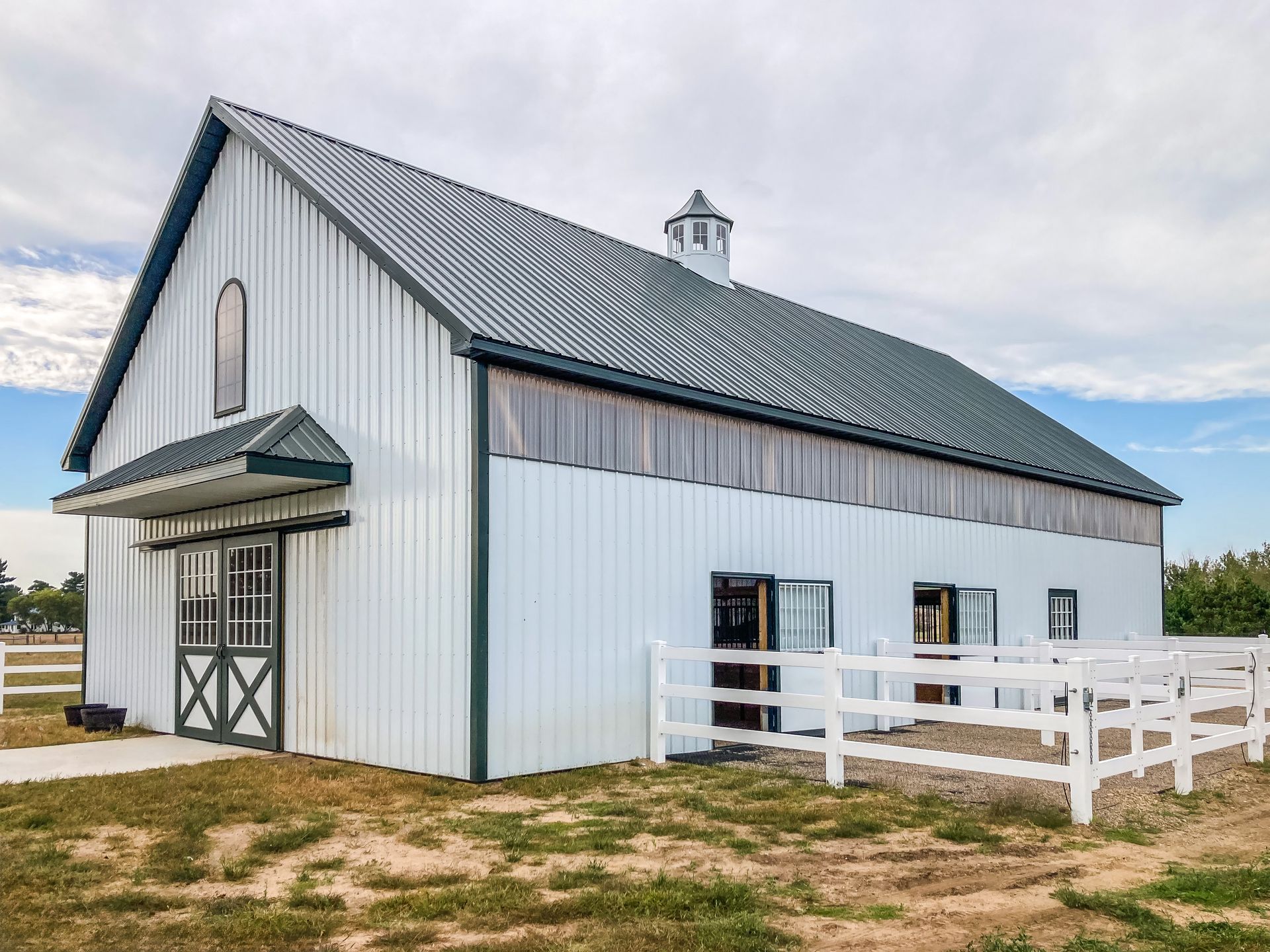 White barn with dark roof, green trim, and white fence on a grassy field under a cloudy sky.