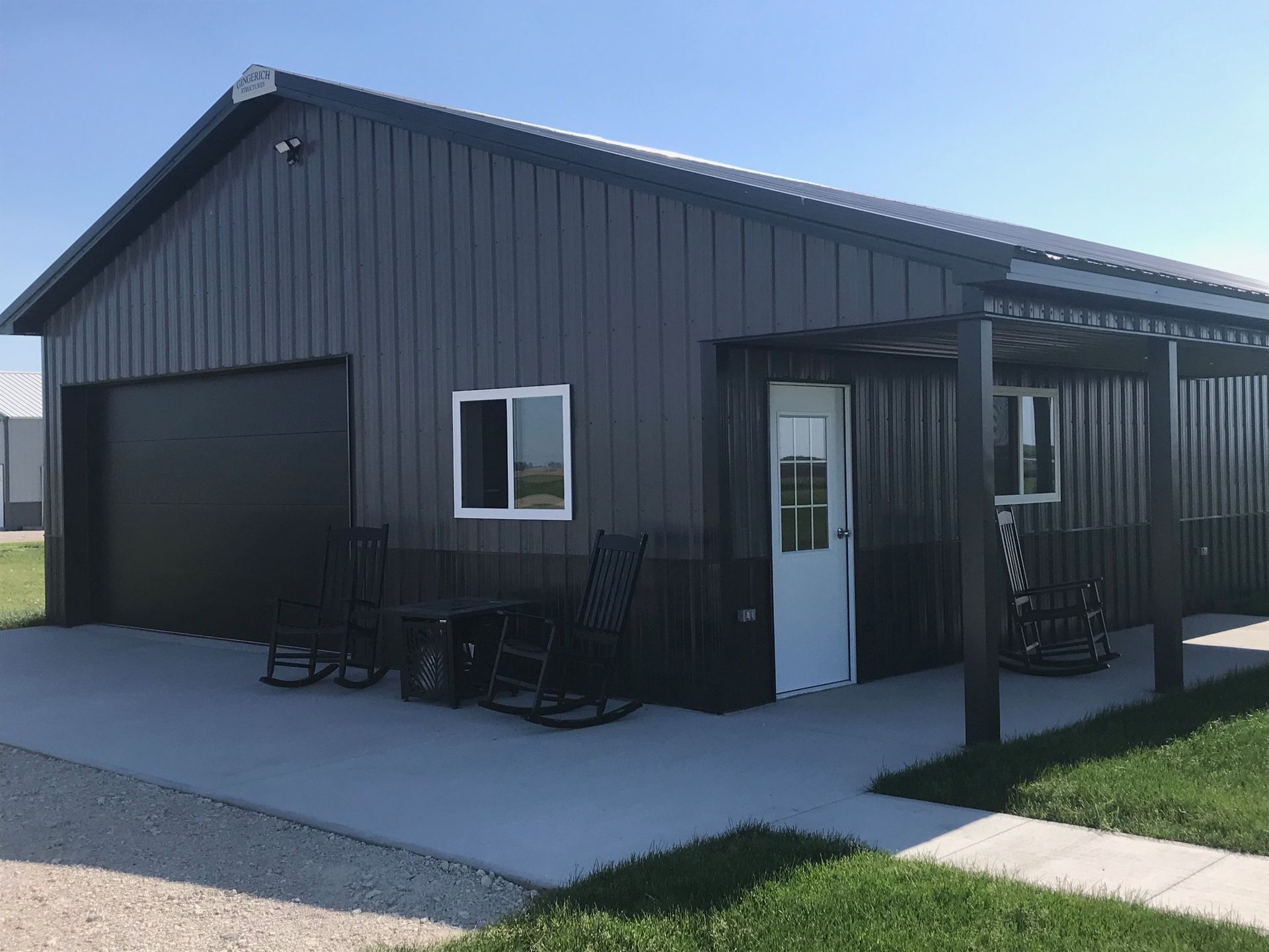 Dark gray metal building with garage door, white door and windows, and a covered porch with rocking chairs.
