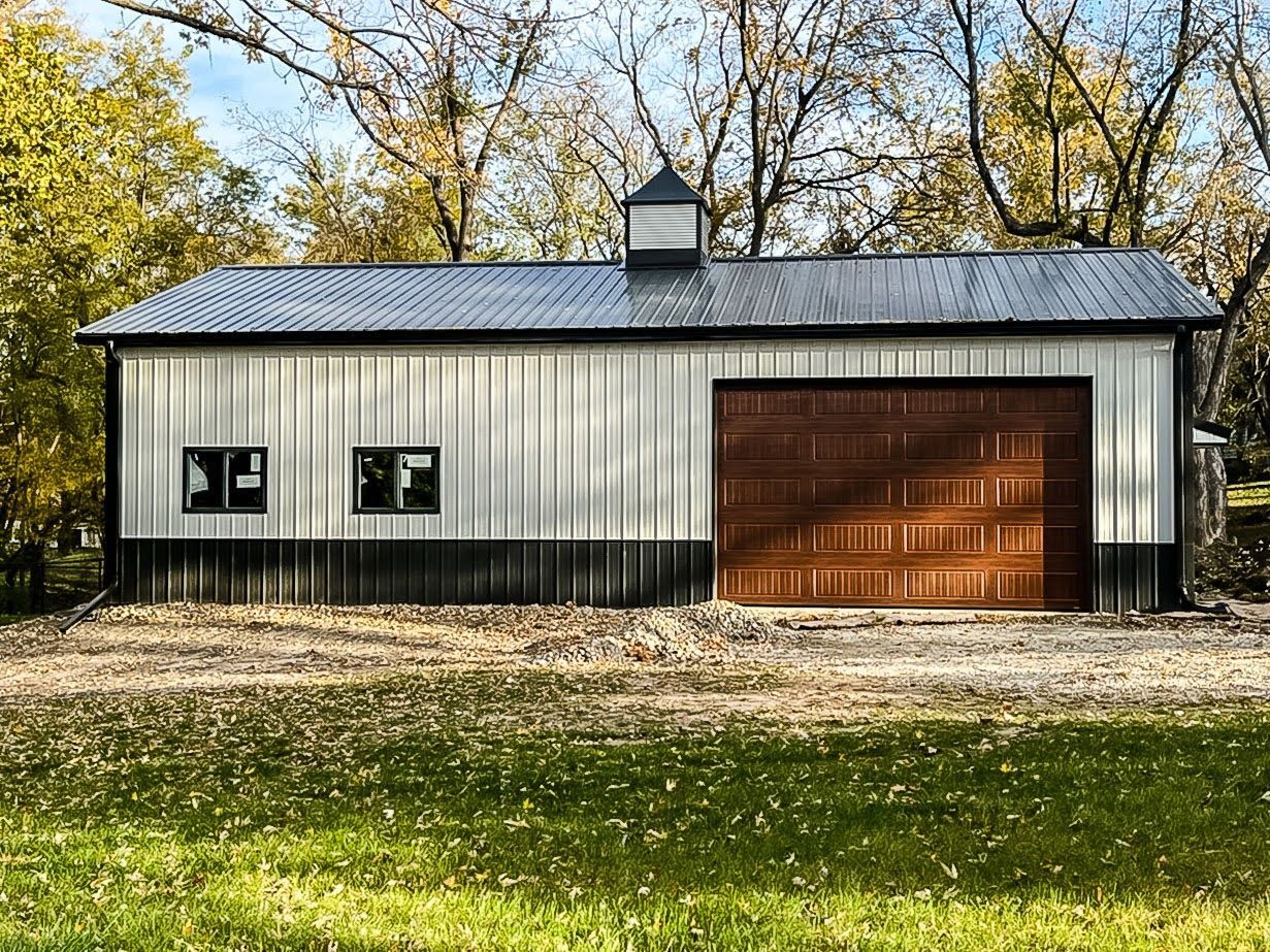 A metal-sided garage with a brown door and black trim, set against fall foliage.