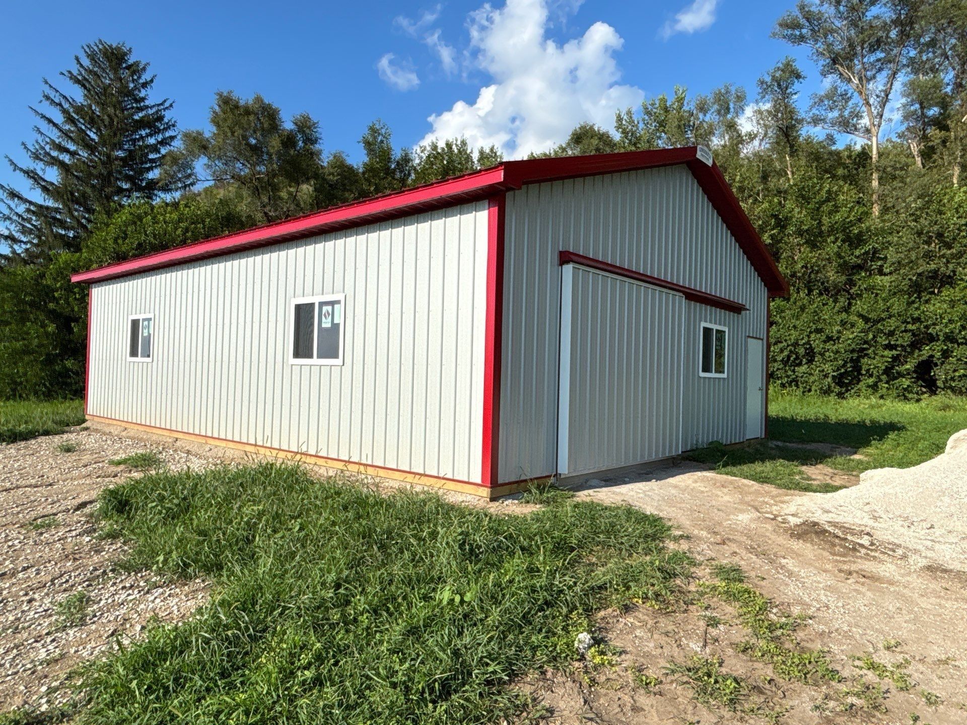 Metal building with red trim, two windows, and open door. Grassy area with gravel driveway.