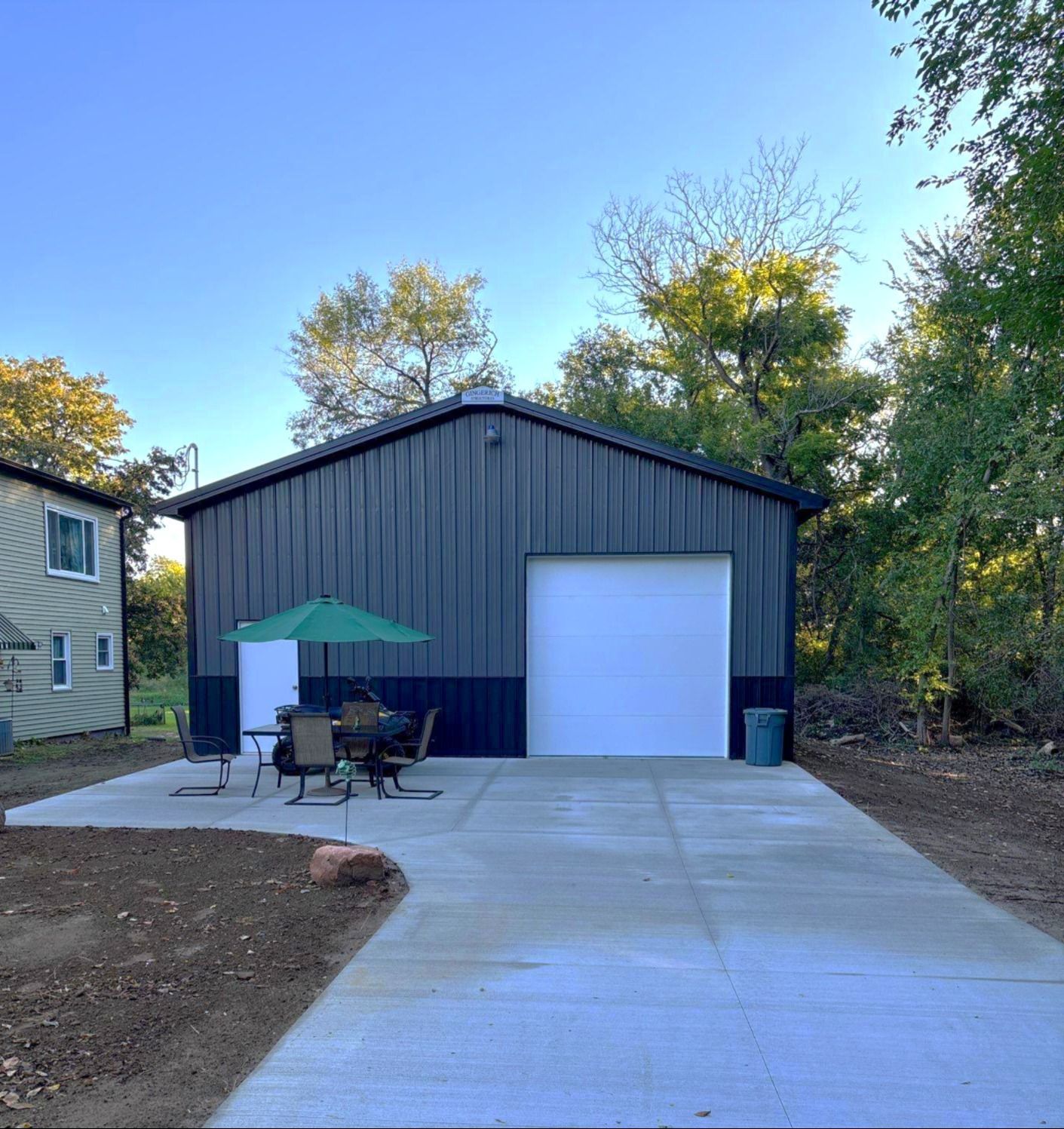 Black and white metal garage with a concrete driveway. An umbrella and seating are on the side. Trees in the background.
