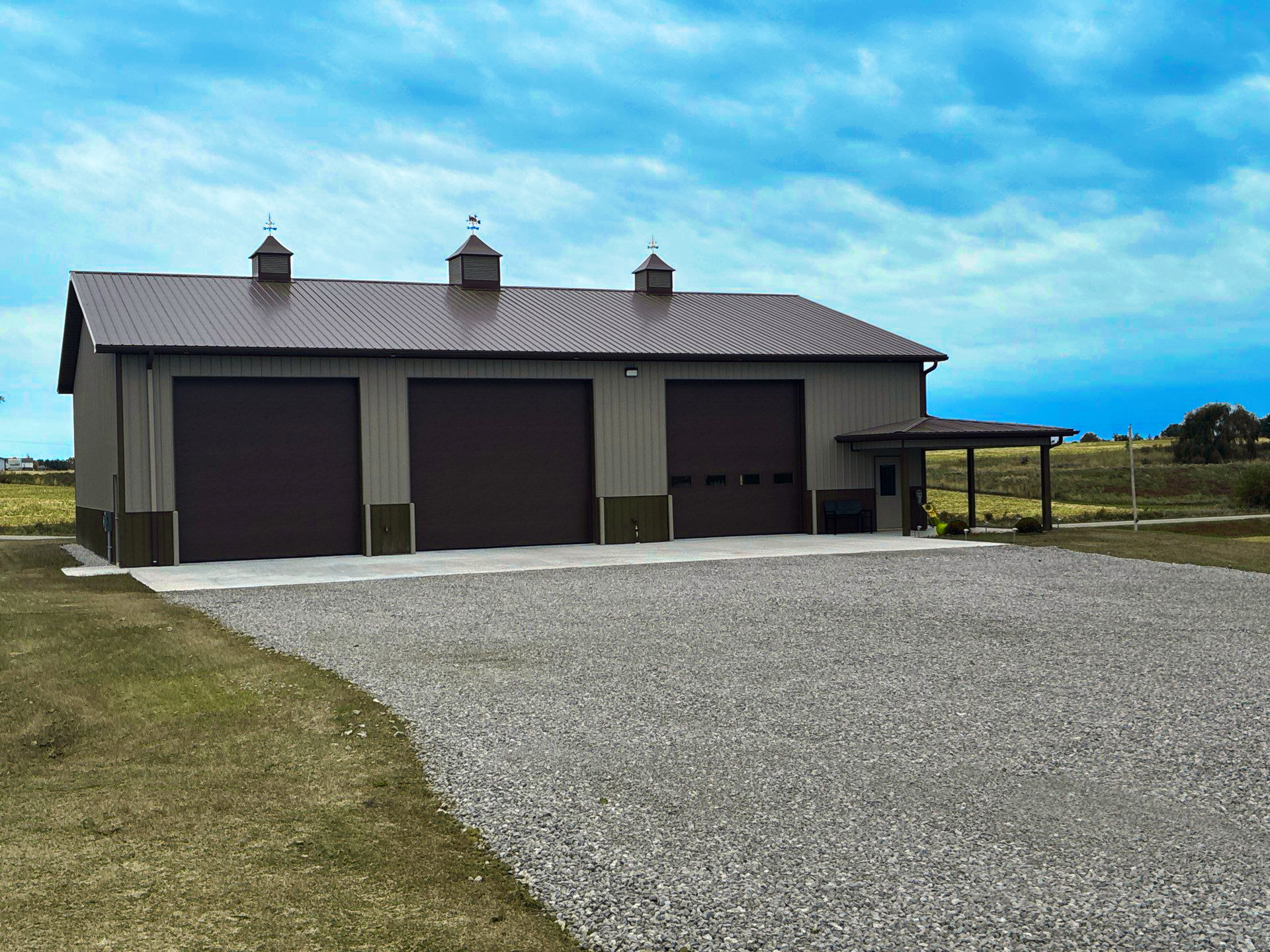Brown and tan metal building with three garage doors and a gravel driveway under a cloudy sky.