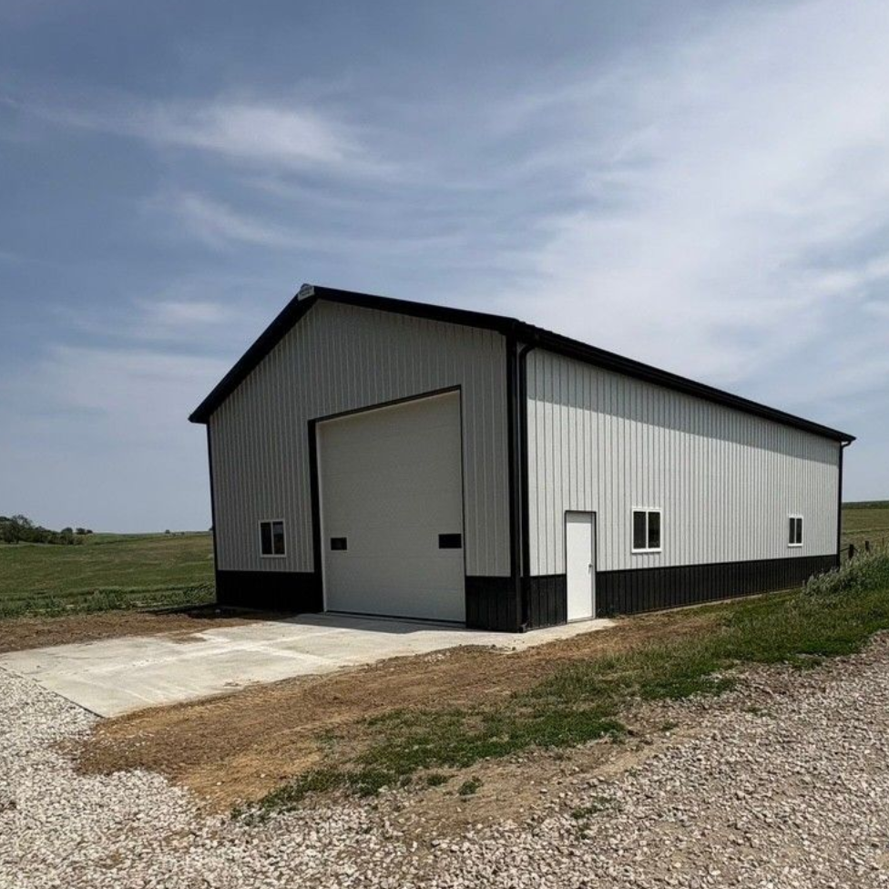 Metal barn with large garage door, white and black siding, set on a gravel driveway under a blue sky.