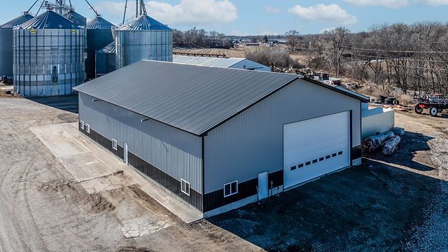 Large gray agricultural warehouse with silos in the background, cloudy sky.