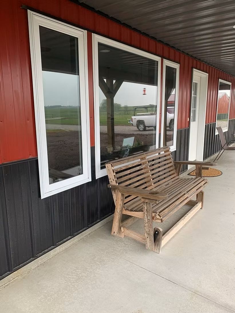Wooden bench on a porch in front of a building with red and black siding and windows.