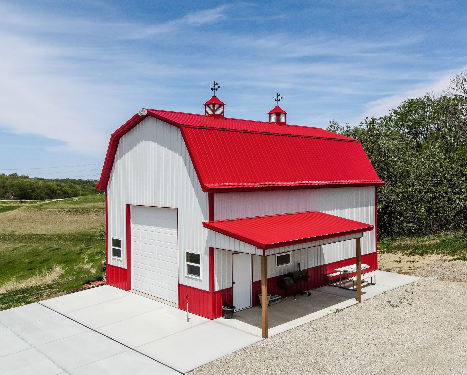 Red and white barn with red roof and small overhang, set on a concrete pad. Two small cupolas. Blue sky.