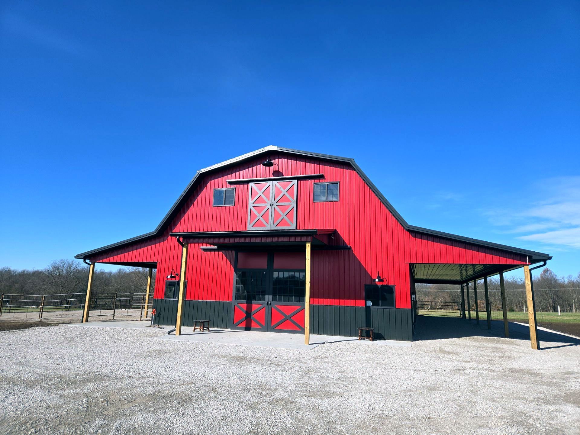 A red barn with dark trim, a gambrel roof, and open side porches, set against a bright blue sky on a gravel lot.