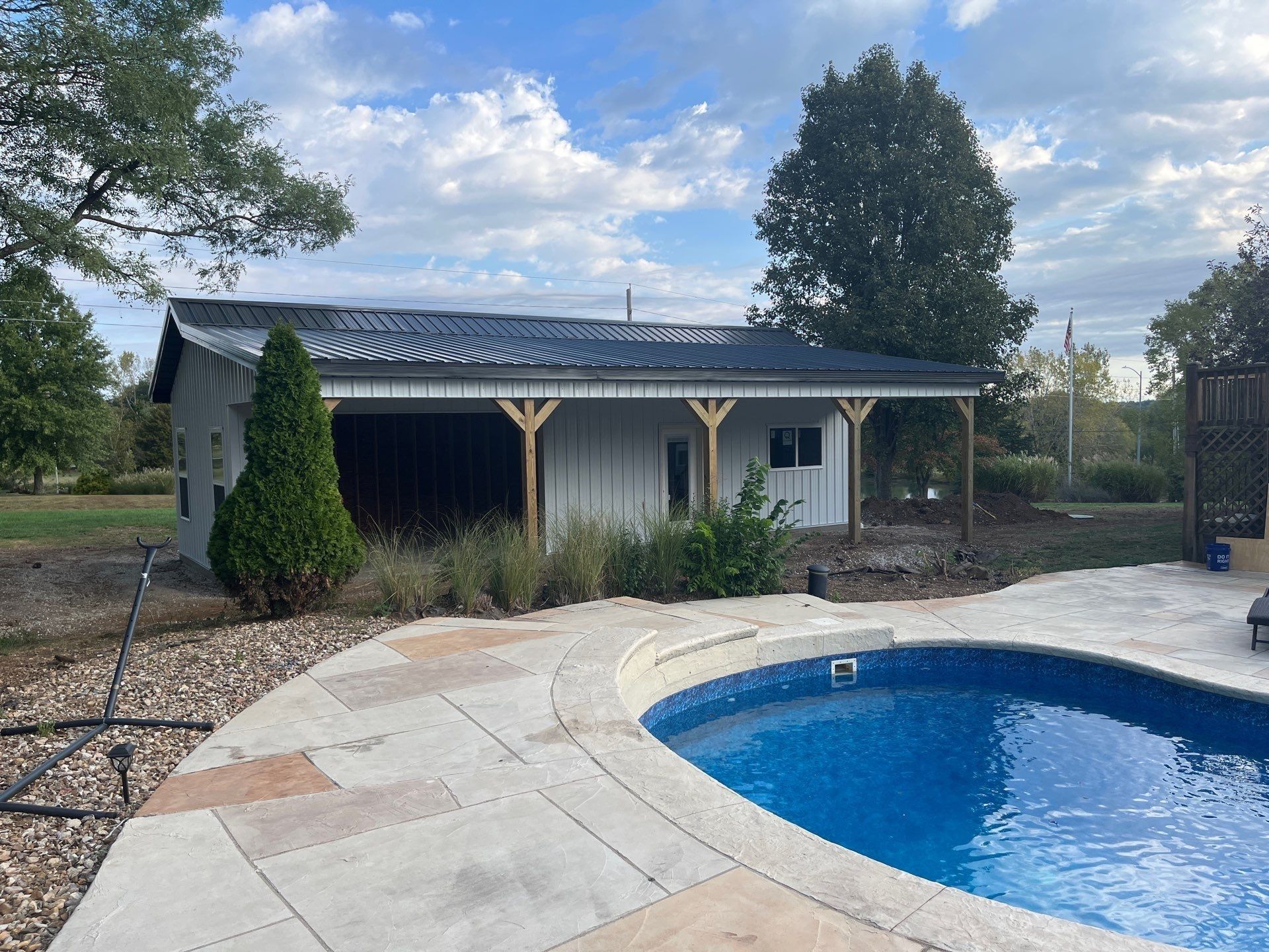 Poolside building with dark roof, white siding, and wooden supports. Blue pool in foreground, grassy yard, cloudy sky.