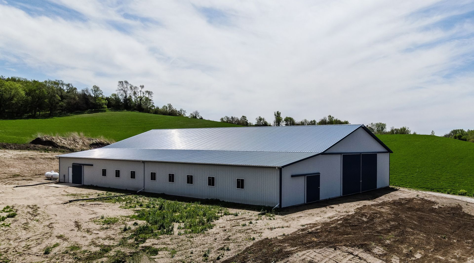 Large white and gray metal barn on a dirt plot with green fields and trees in the background.