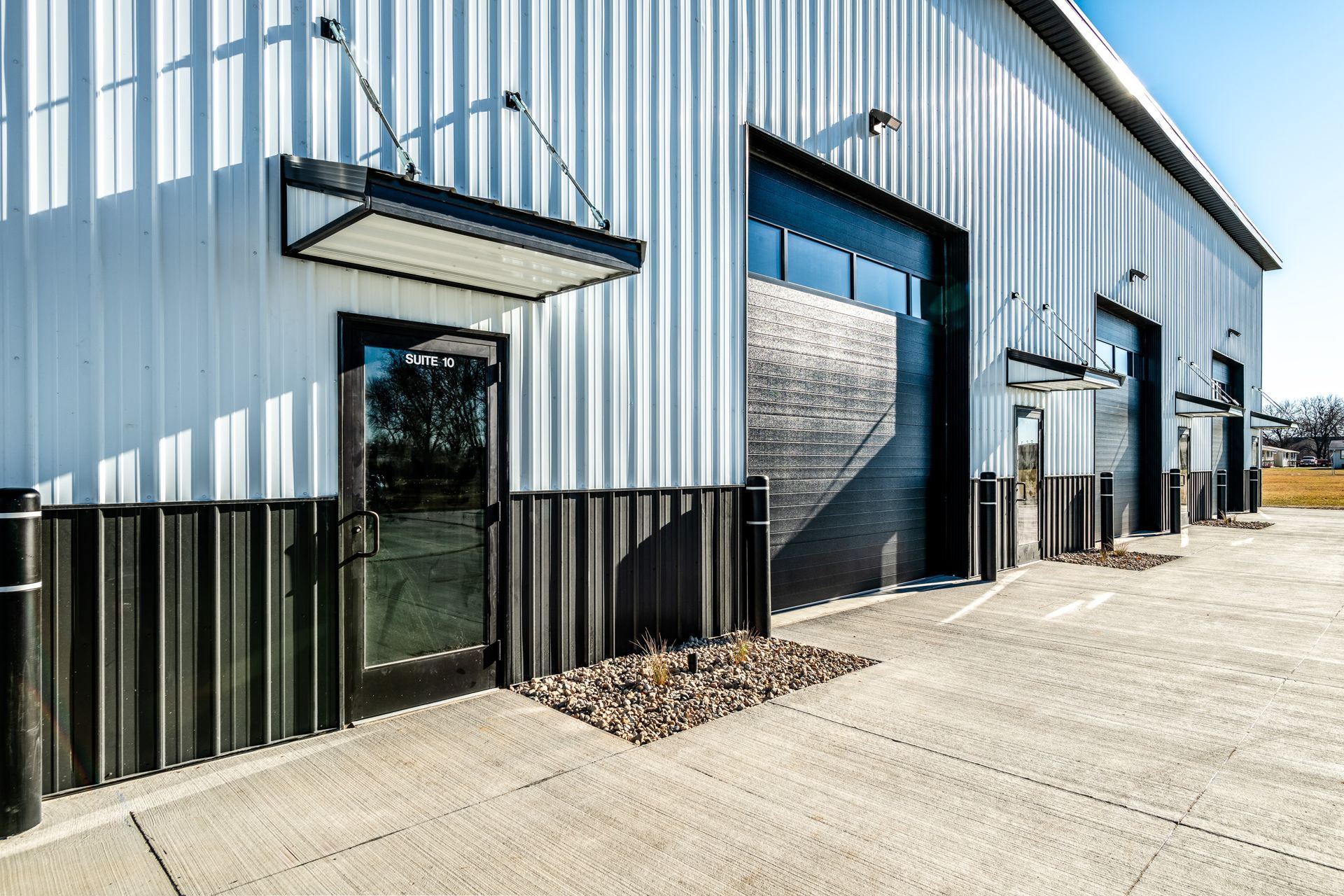 White and black industrial building with a glass door and garage doors on a concrete lot.