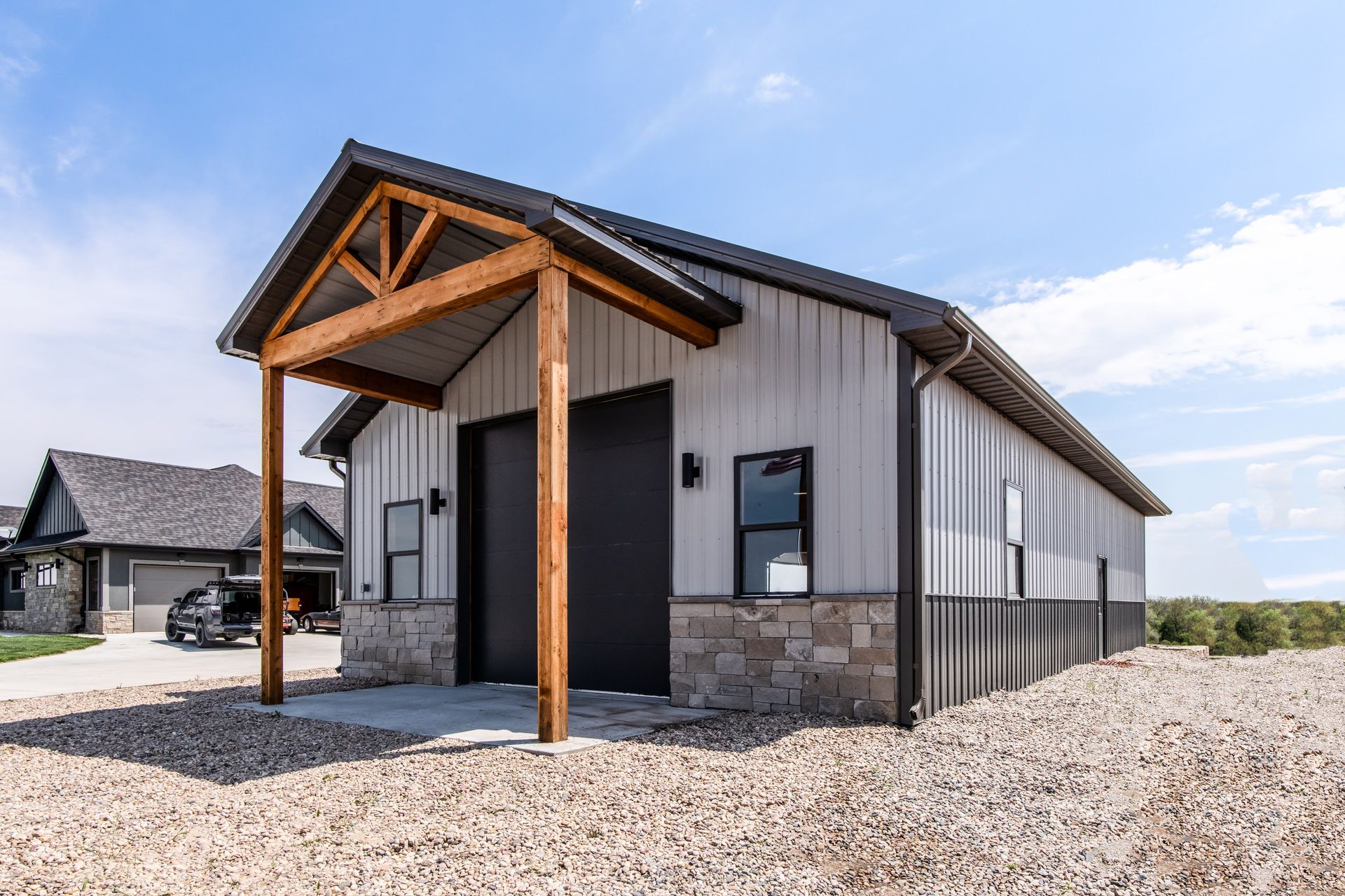 Metal-sided building with stone base and wood porch. Includes black garage doors, windows, and a gravel driveway.