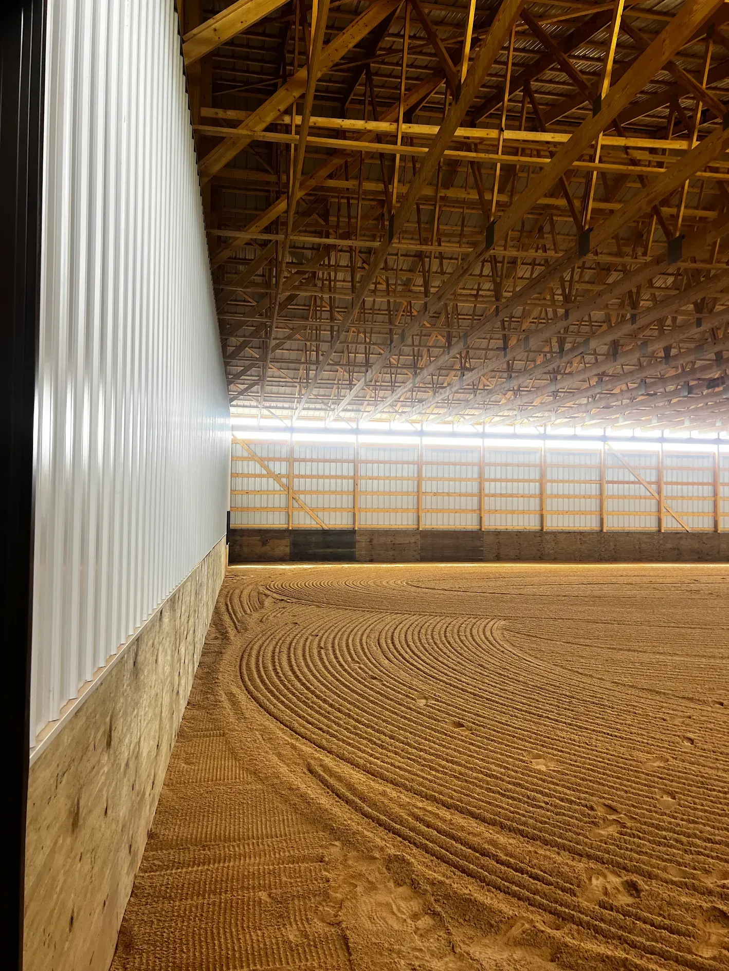 An indoor horse arena with sand footing, corrugated metal walls, and wooden beams.
