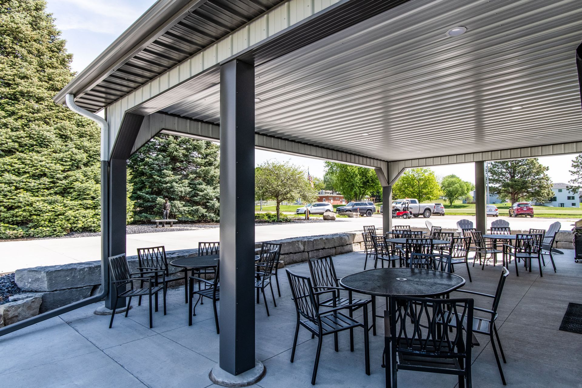 Outdoor patio with tables and chairs under a covered area; cars and trees visible in the background.