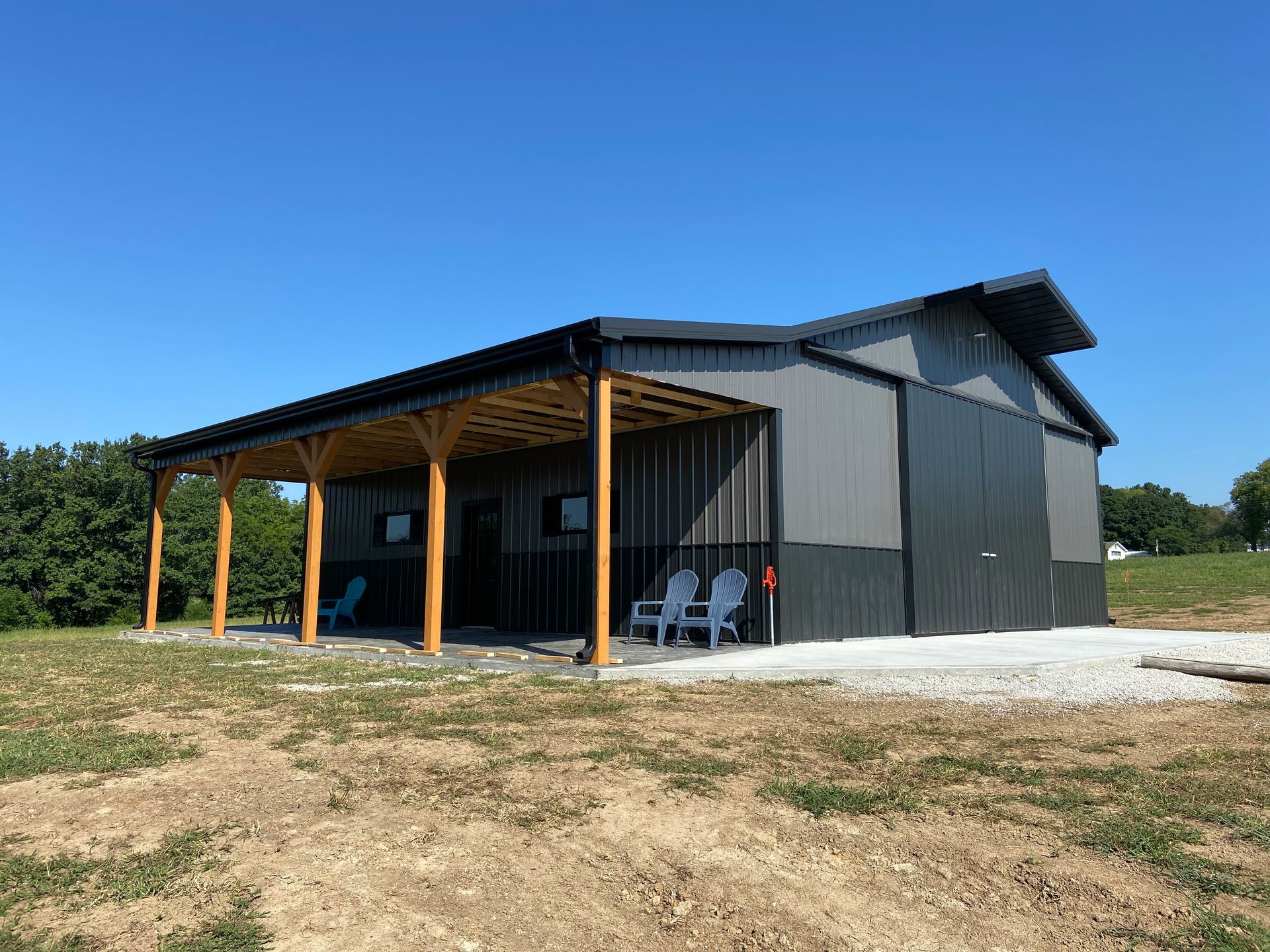 A two-toned gray metal barn with a wooden porch and blue chairs on a sunny day.