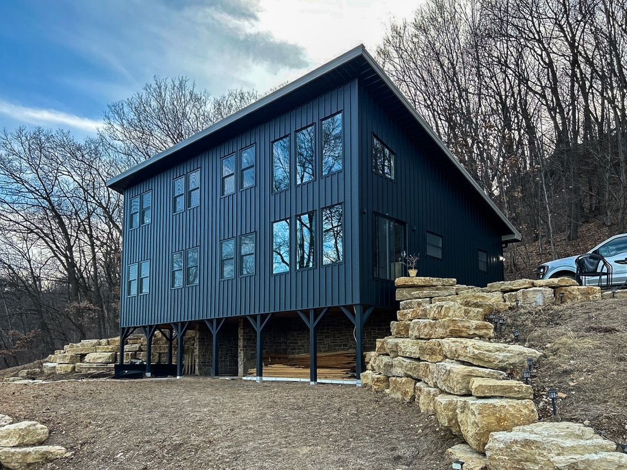 Black modern cabin on raised supports, with stone retaining wall, set in a wooded area.