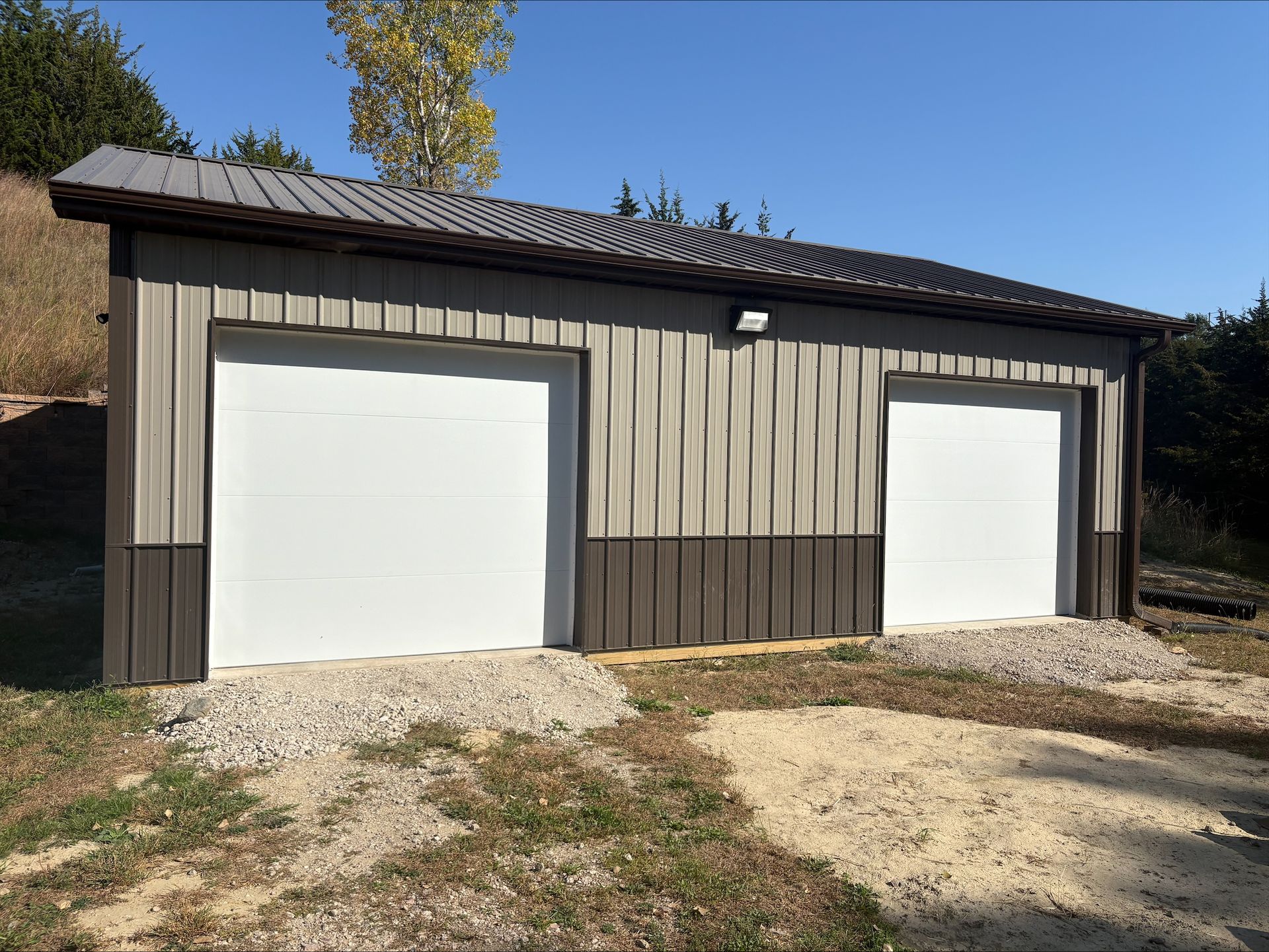 Tan and brown metal garage with two white garage doors, set on gravel.