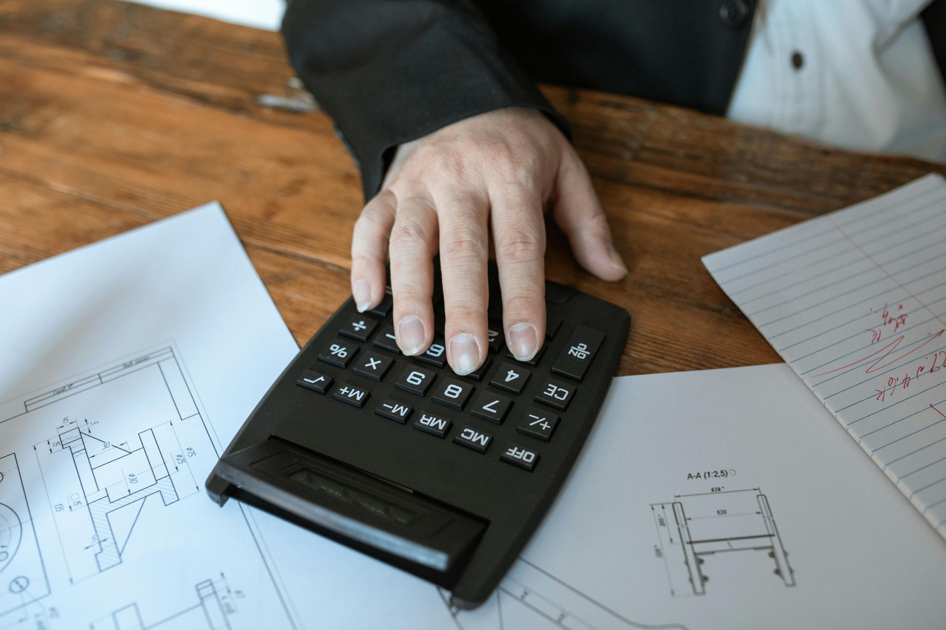 Person's hand on a black calculator, surrounded by papers with diagrams on a wooden desk.
