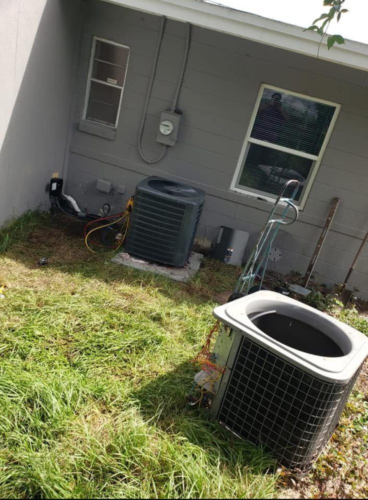 Two air conditioning units in grassy yard next to gray building with windows and electrical box.