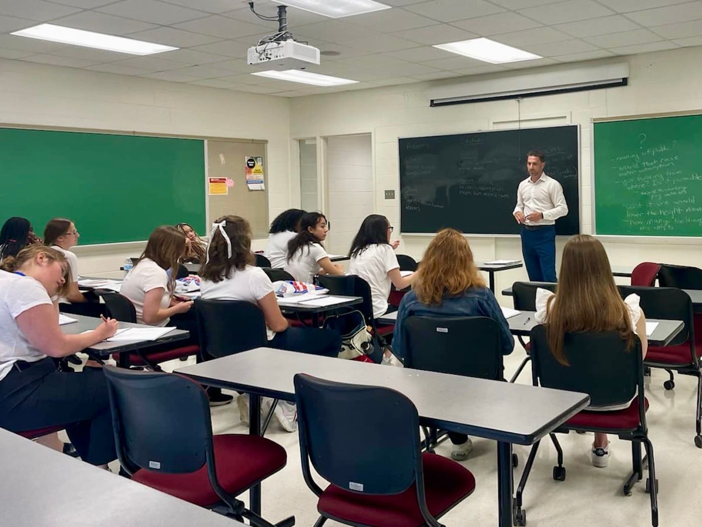 A group of young women are sitting at desks in a classroom with a man standing before them