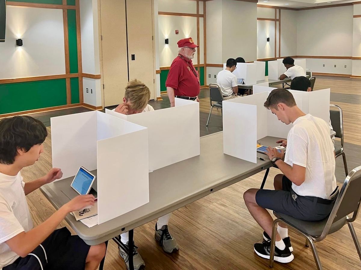 A group of young men are sitting at tables with dividers between them as they cast voting ballots on tablet computers