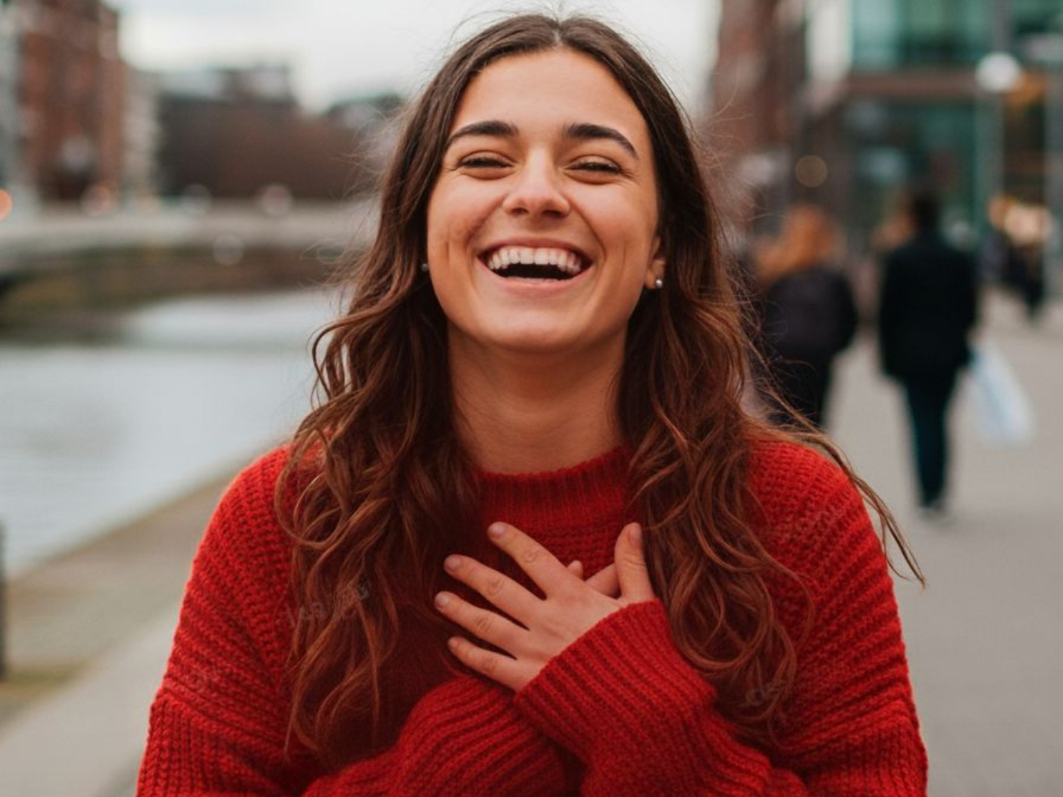 Une femme en pull rouge rit, les mains sur la poitrine, à l'extérieur, près d'un cours d'eau et de bâtiments.